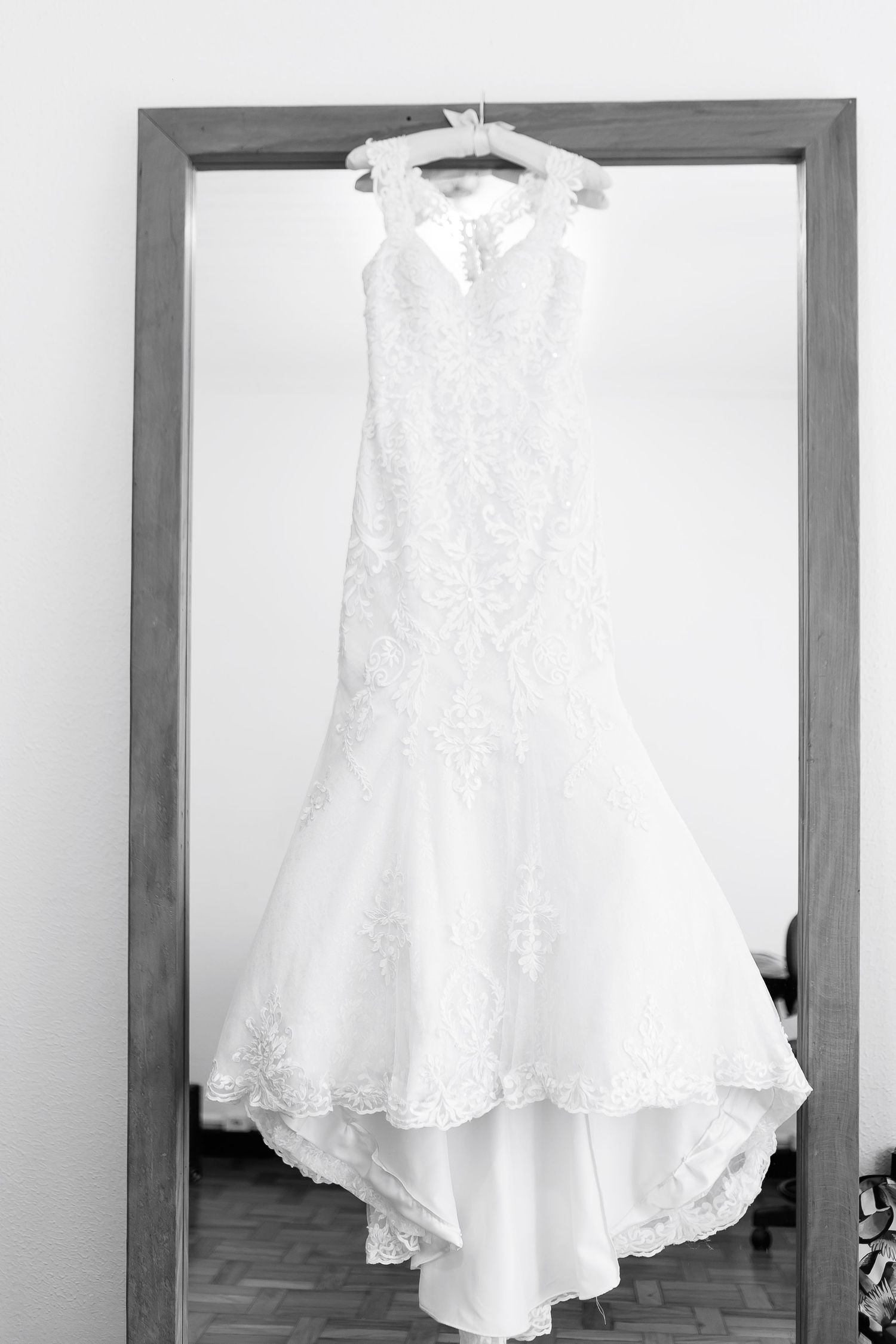 Black and white photo of an elegant lace mermaid wedding dress hanging on a mirror frame.