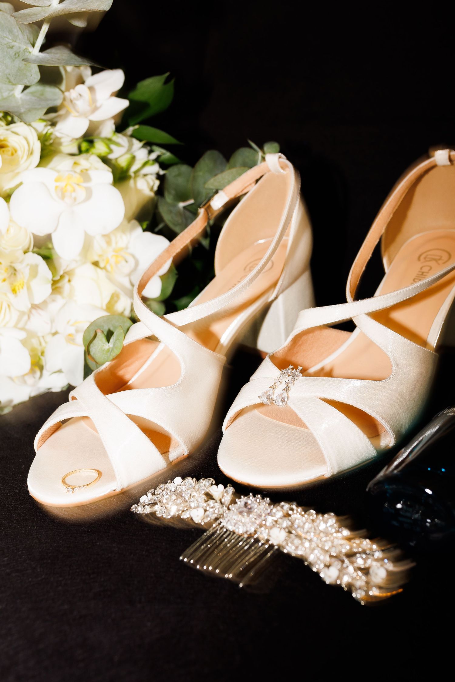 Elegant white bridal heels with pearl hair comb and white floral bouquet on dark background.