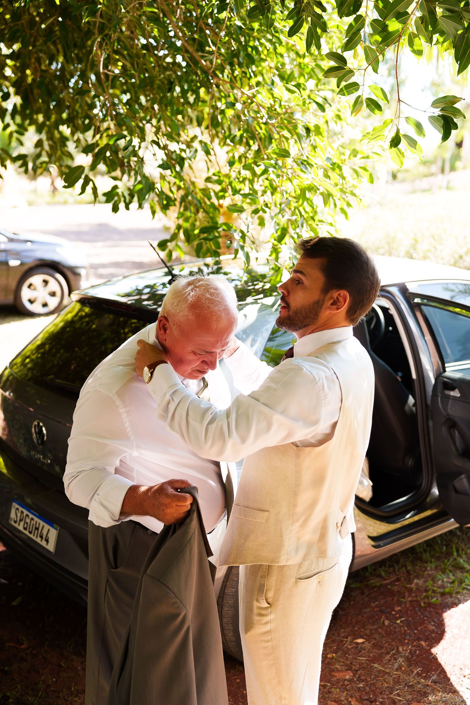 Groom and older man in white shirts adjusting attire outdoors near black car before wedding ceremony.