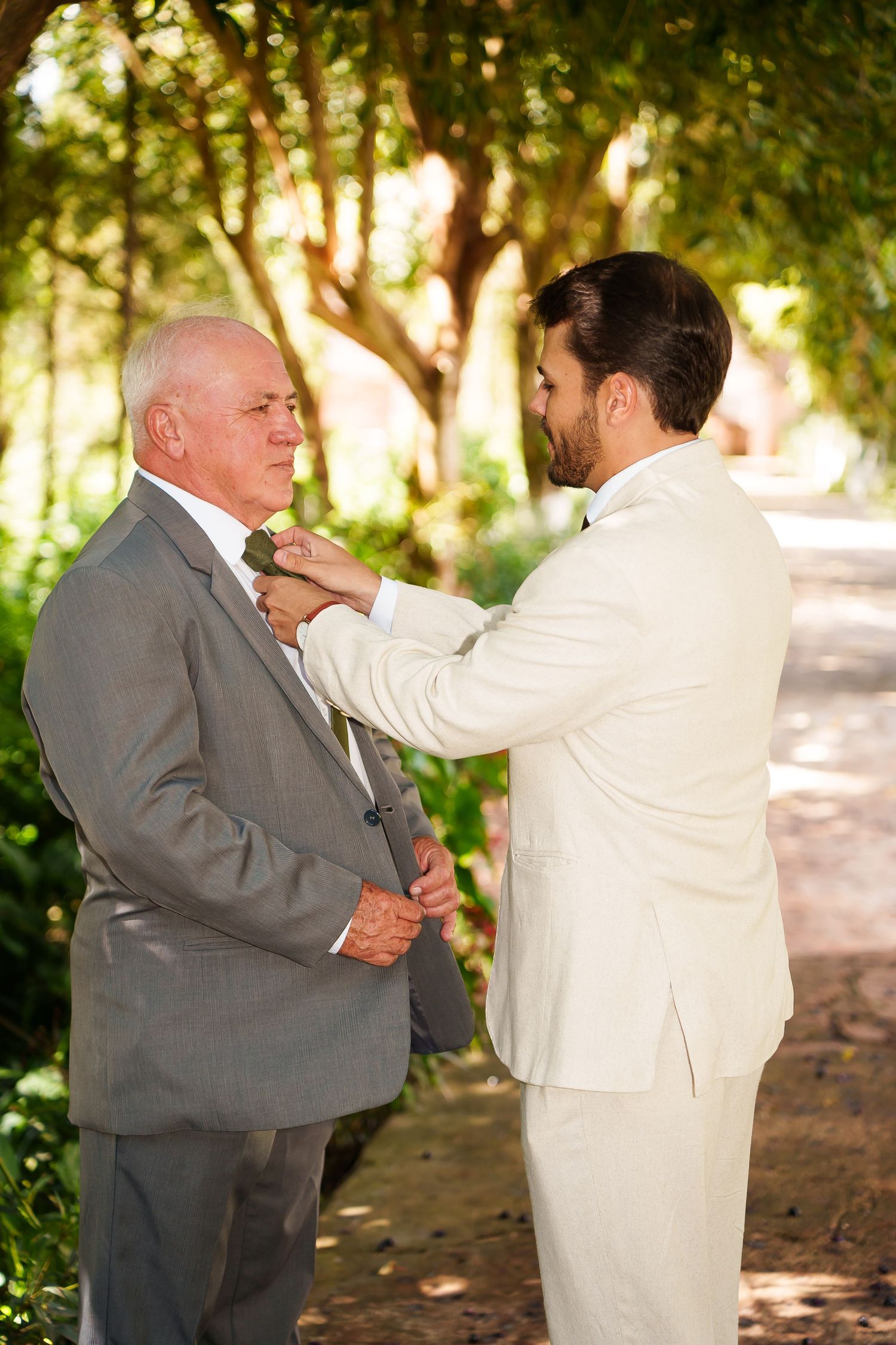 Groom in white suit pins boutonniere on older man in gray suit outdoors at wedding ceremony.