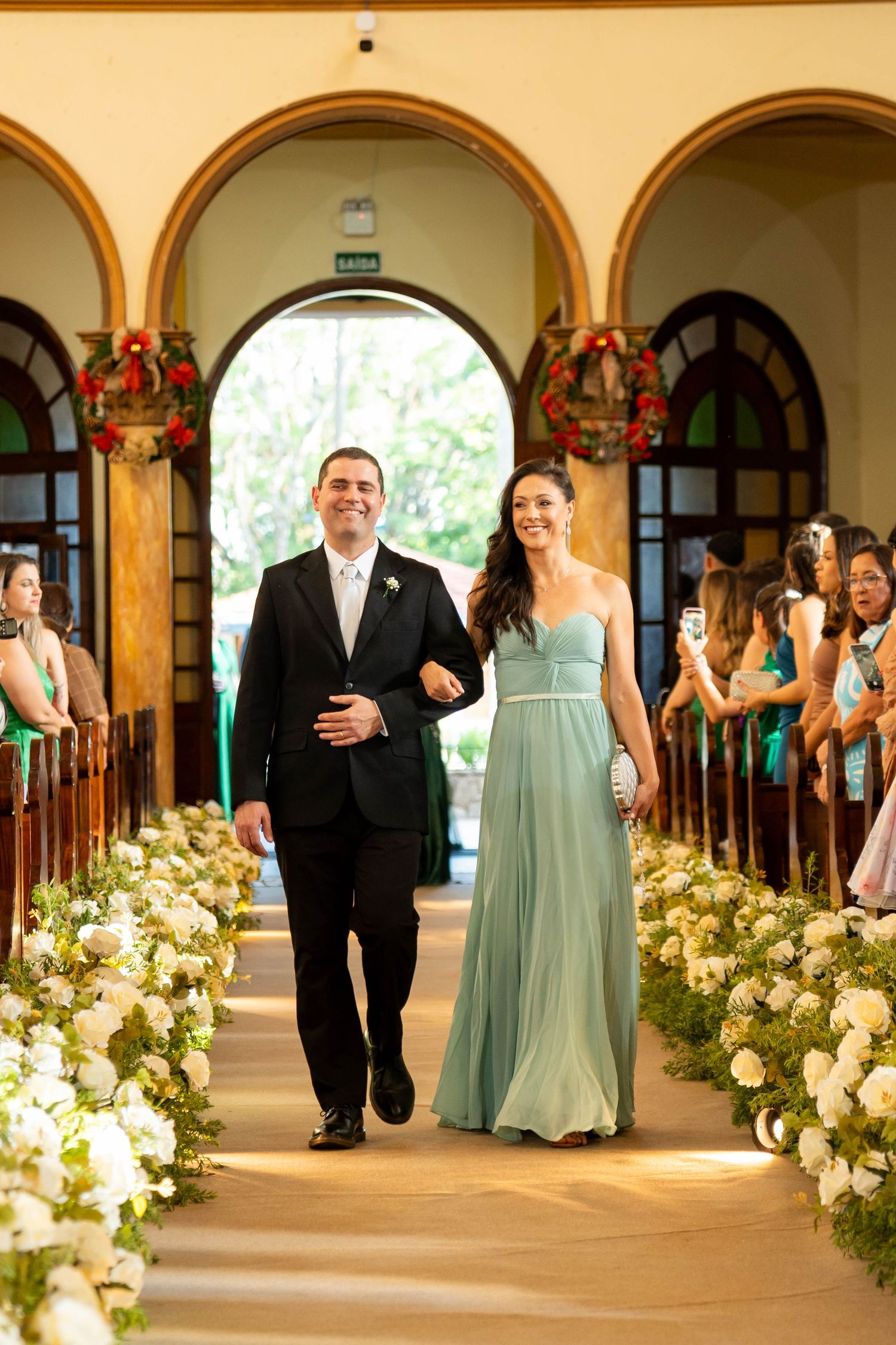A couple walks down a flower-lined church aisle, man in black suit, woman in teal gown, during a wedding ceremony.