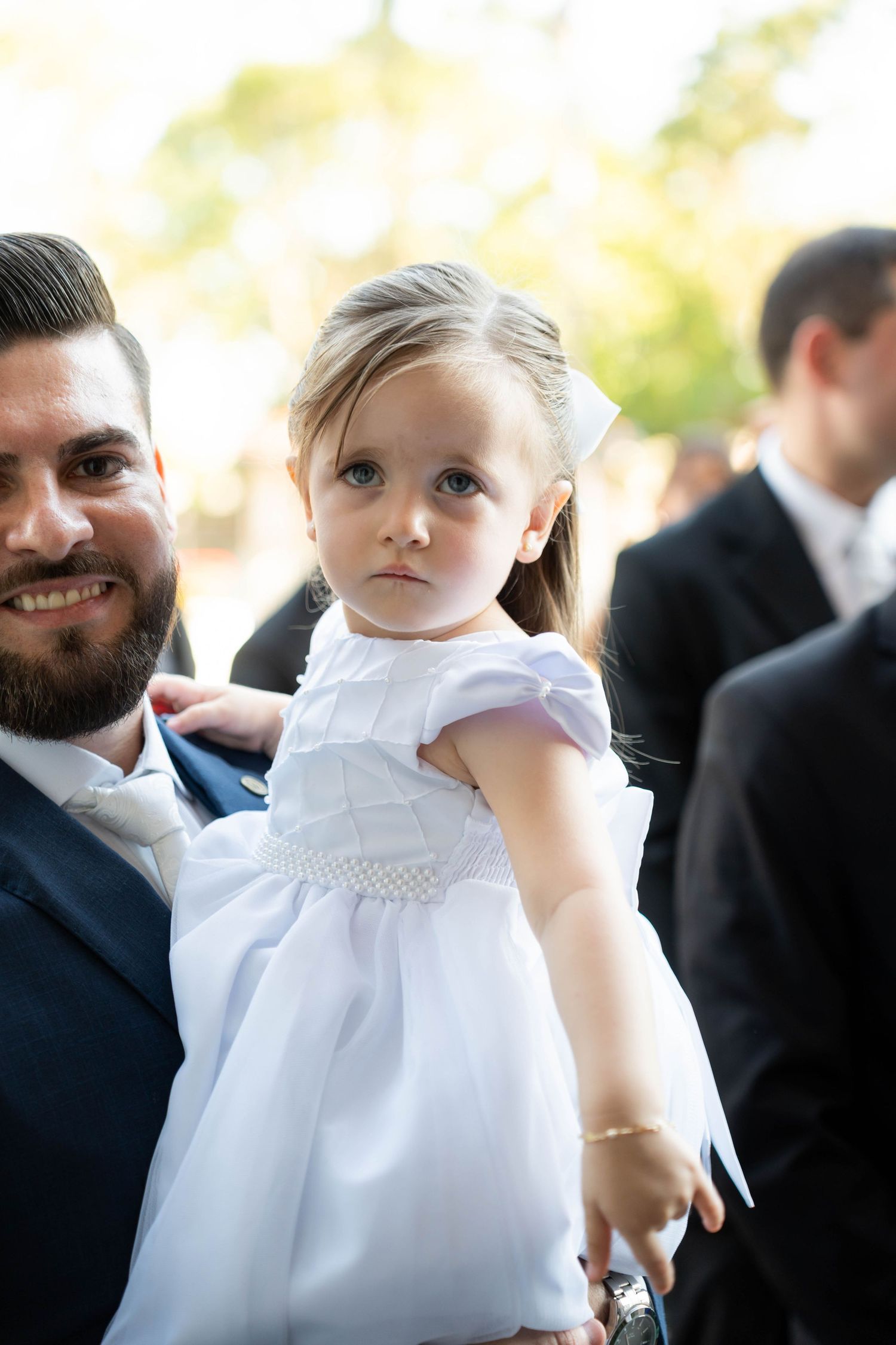 Young flower girl in white dress held by bearded man in navy suit at outdoor wedding ceremony.