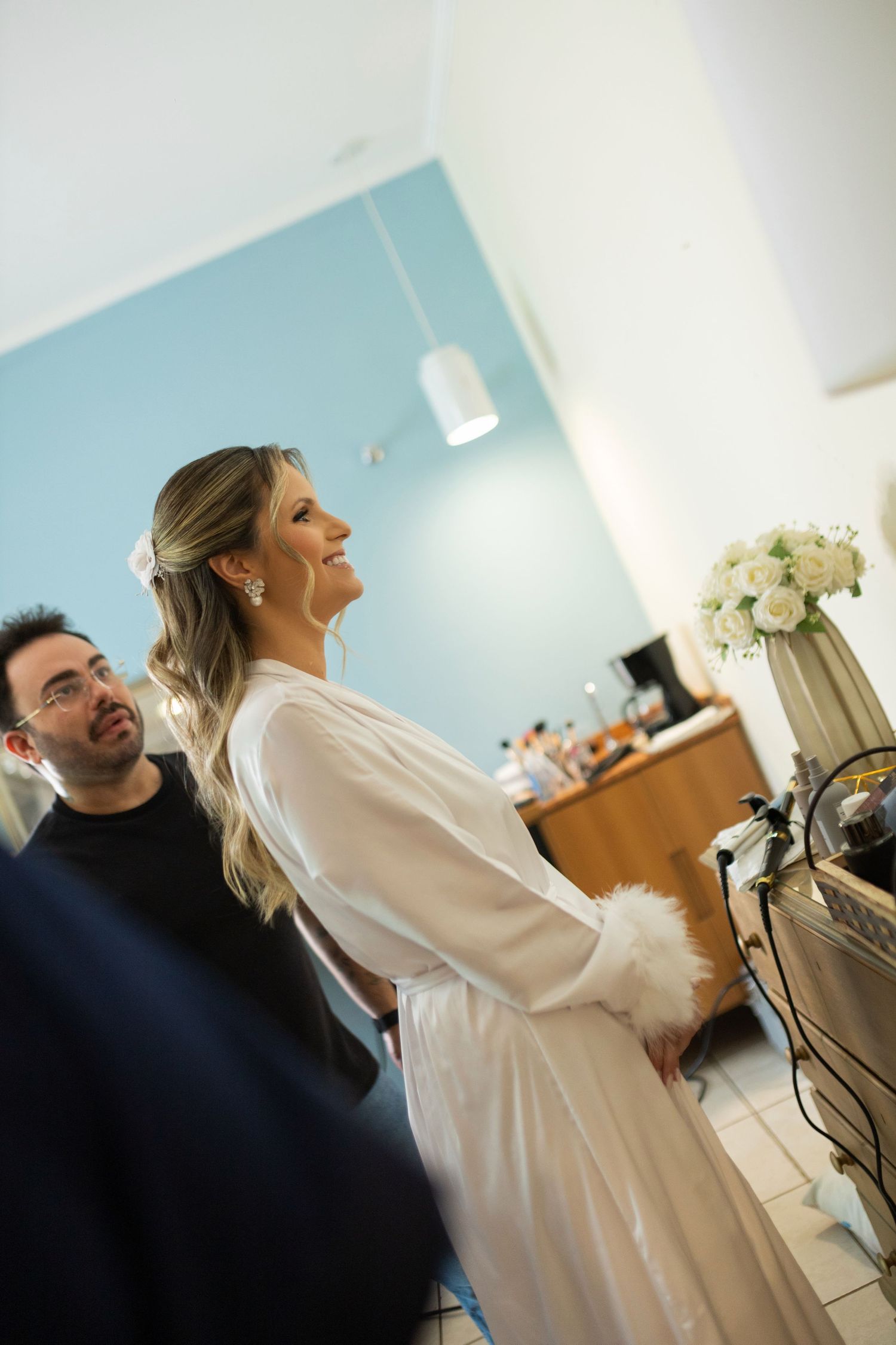 Bride in white robe smiling with hair stylist in a bright salon with floral arrangement nearby.