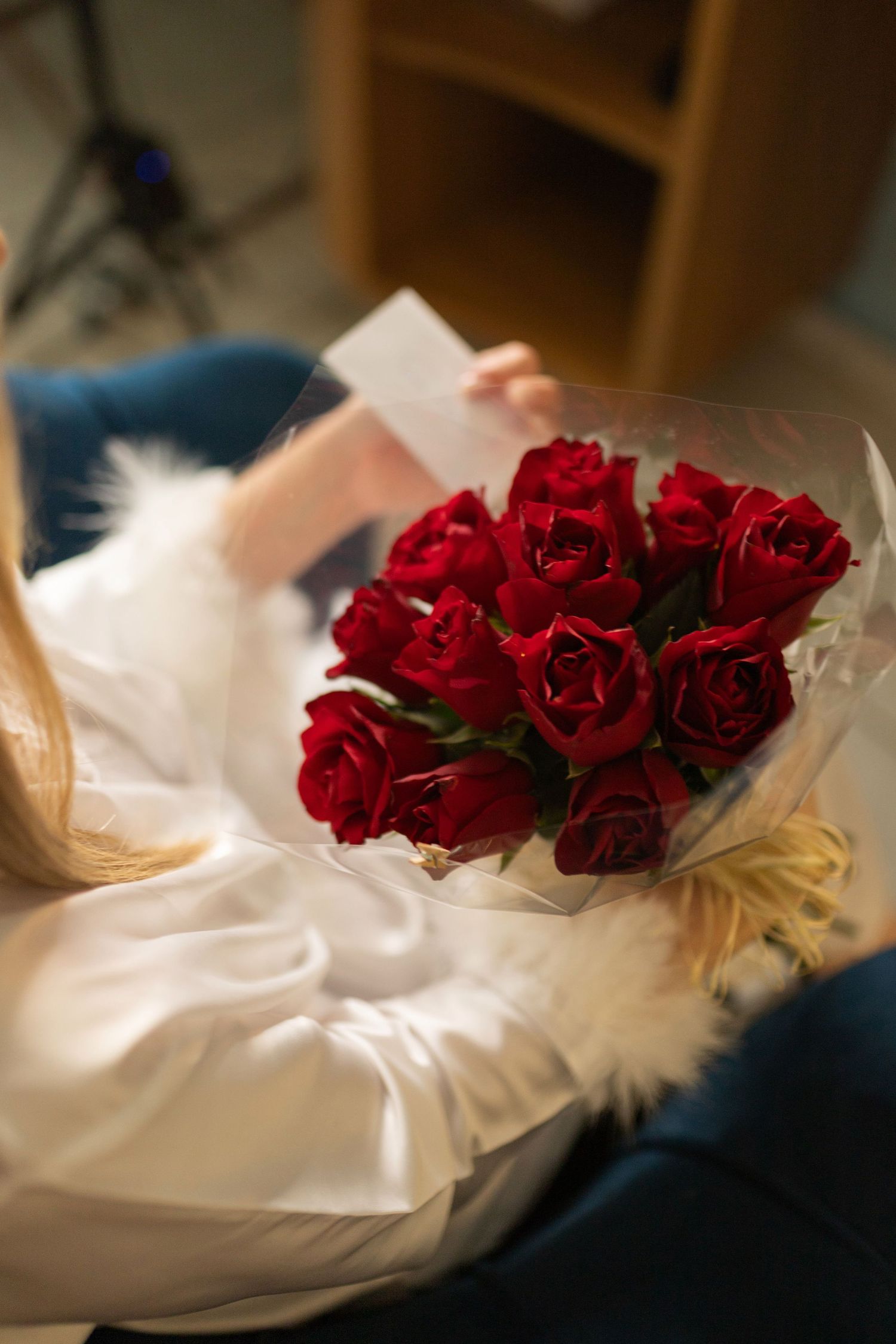 Woman in white robe holding red roses bouquet wrapped in white paper, reading a card.
