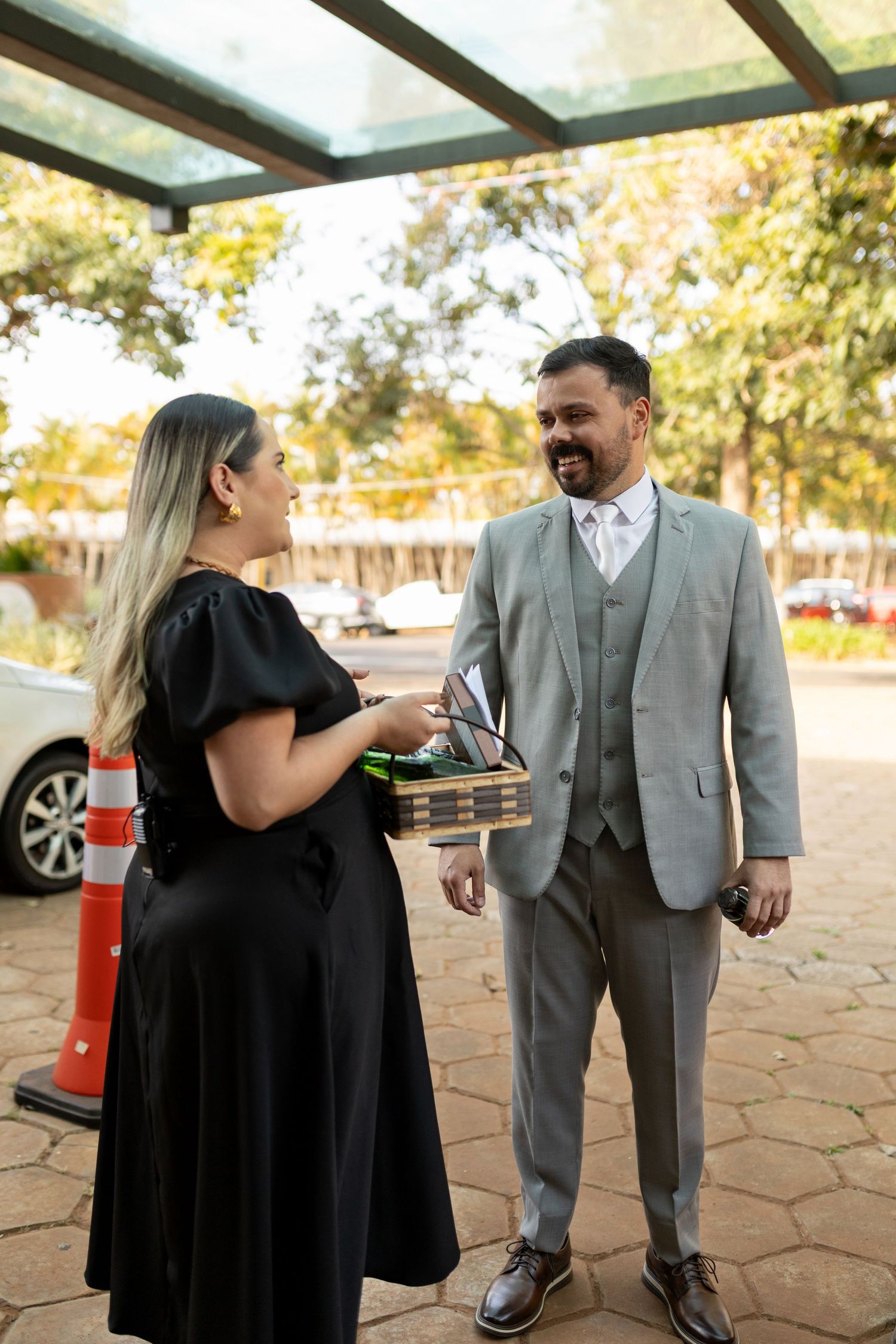 A woman in black dress hands items to a smiling man in gray suit outdoors near a covered entrance.