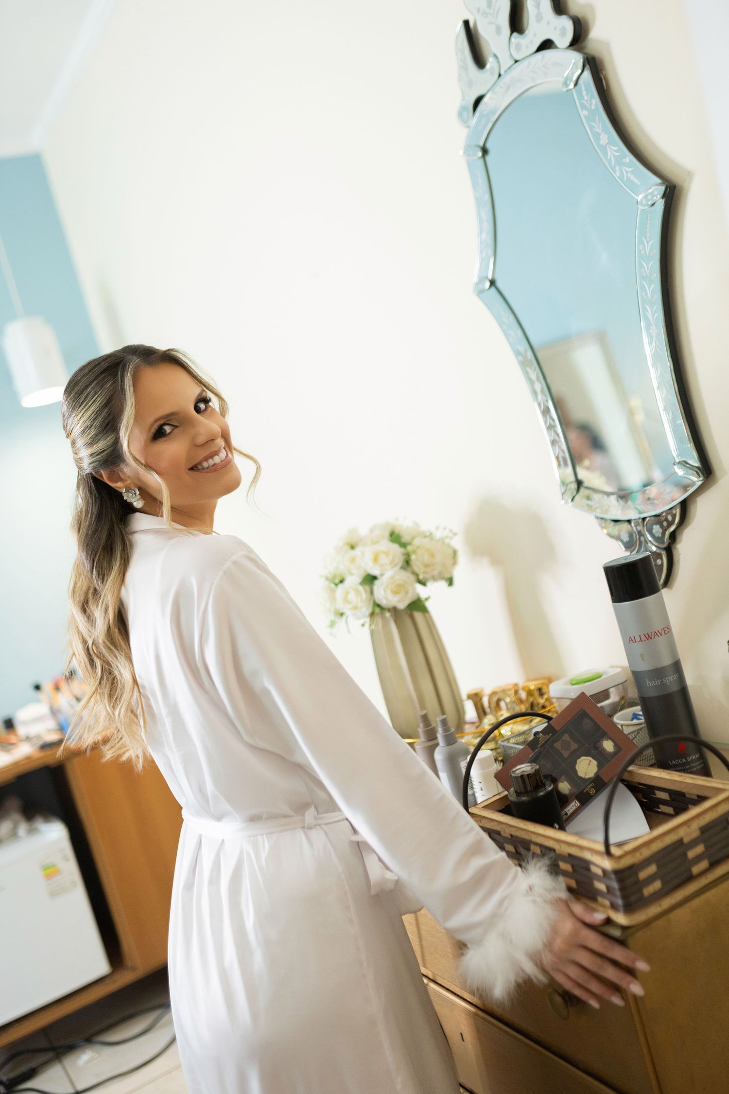 Smiling bride in white robe stands at vanity with ornate mirror and white flowers while getting ready.