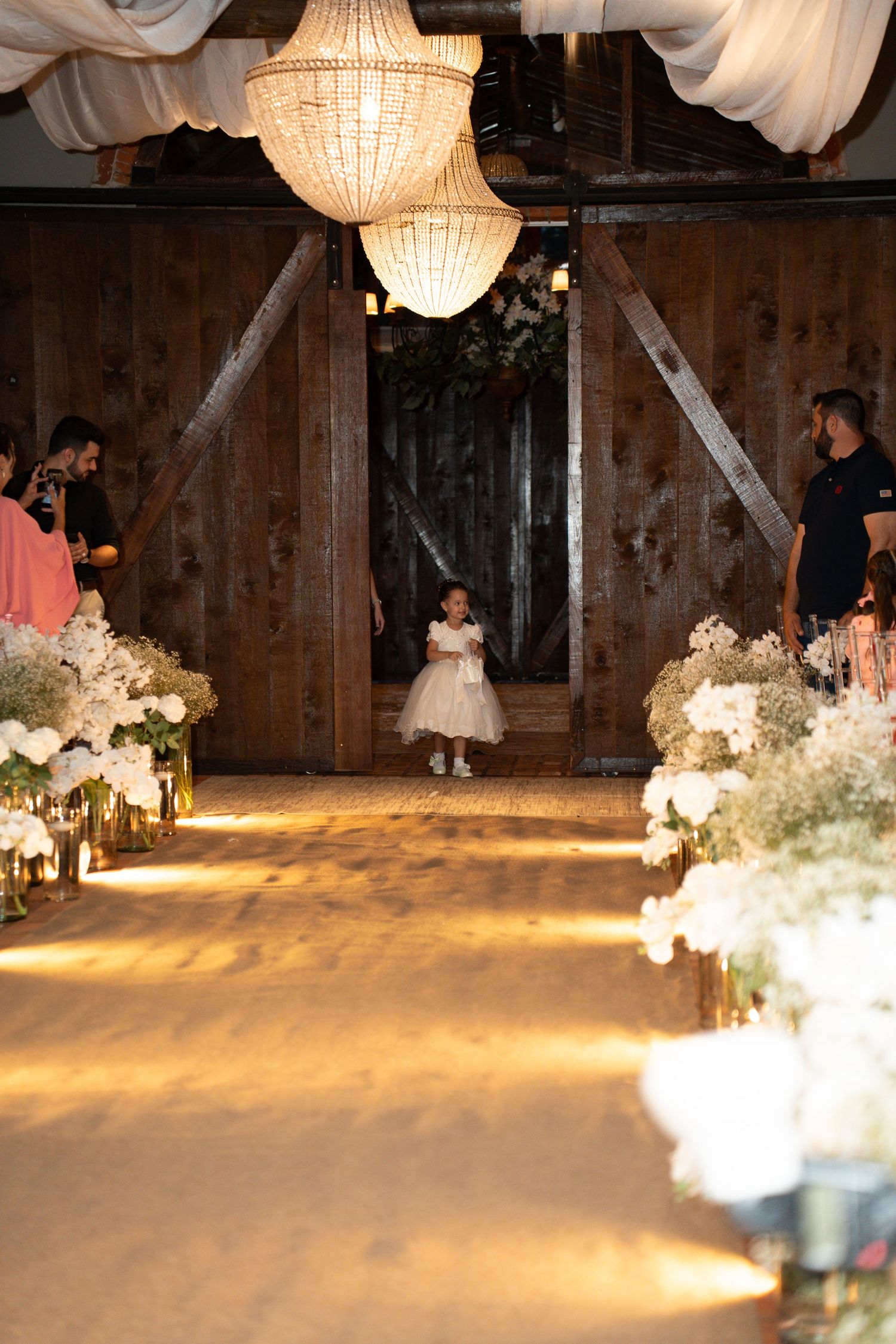 A flower girl in a white dress walks down a floral-lined wedding aisle in a rustic barn venue with chandeliers.