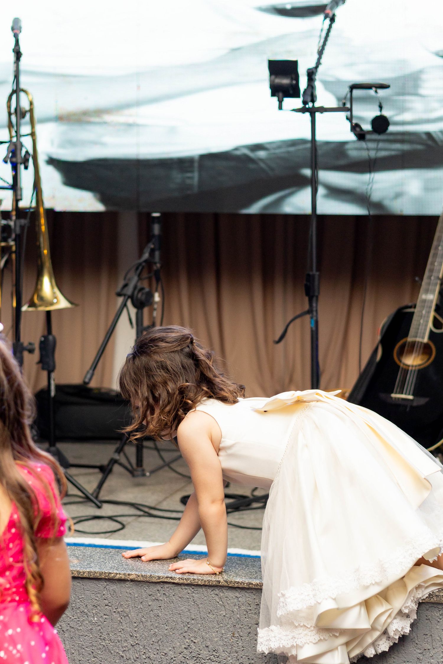Bride in white dress bending down on stage near musical instruments including guitar and trombone.