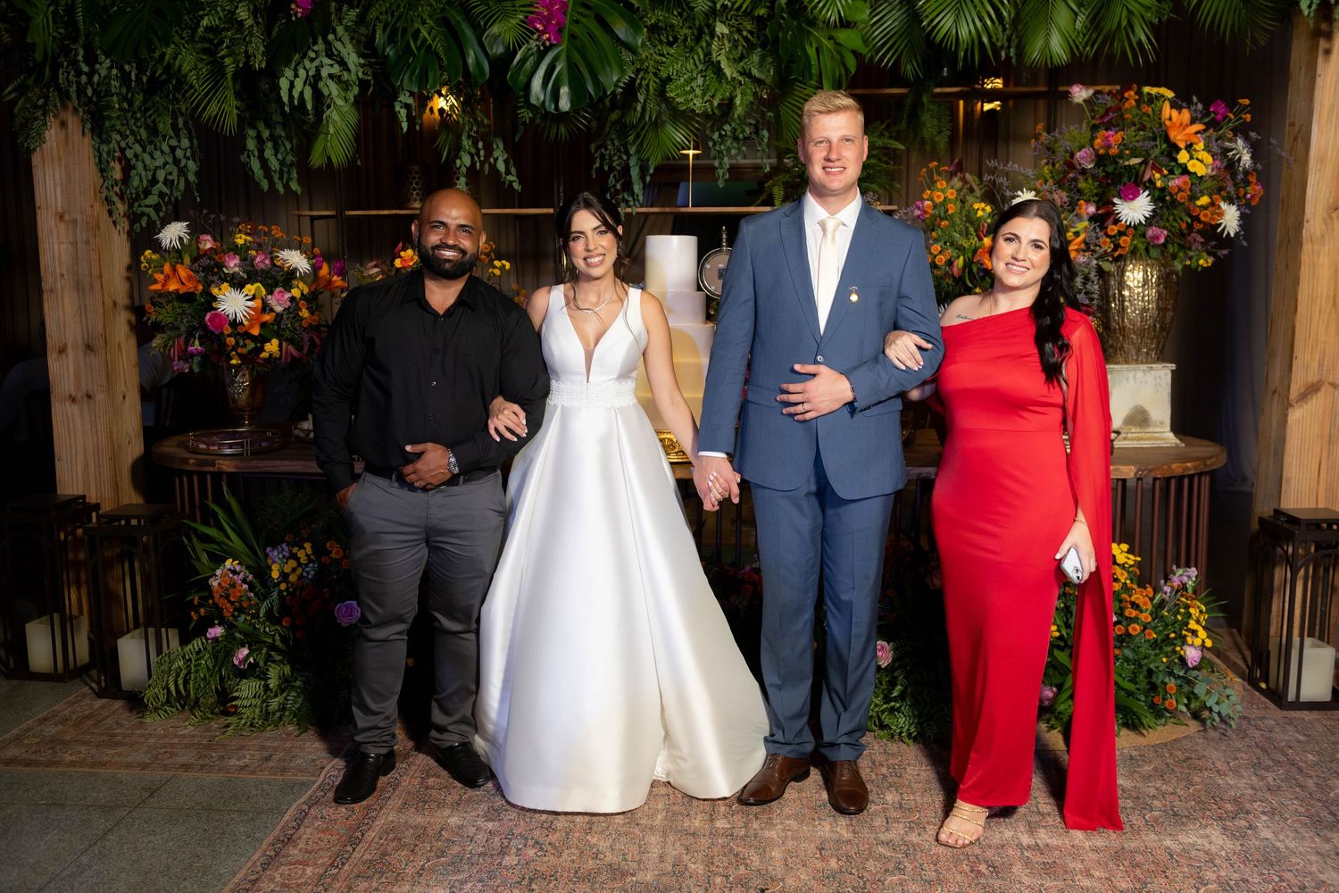 Two couples pose together at a wedding, one bride in white gown, groom in blue suit, and guests in formal attire.