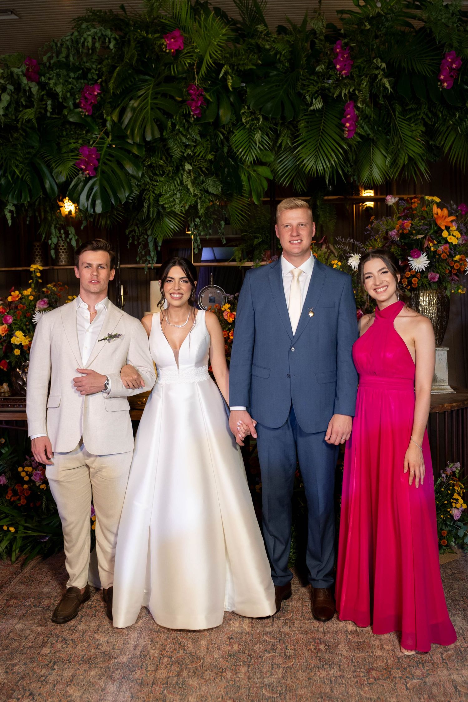 Wedding party of four posing before tropical floral backdrop, bride in white gown, groom in blue suit, guests in pink and beige.