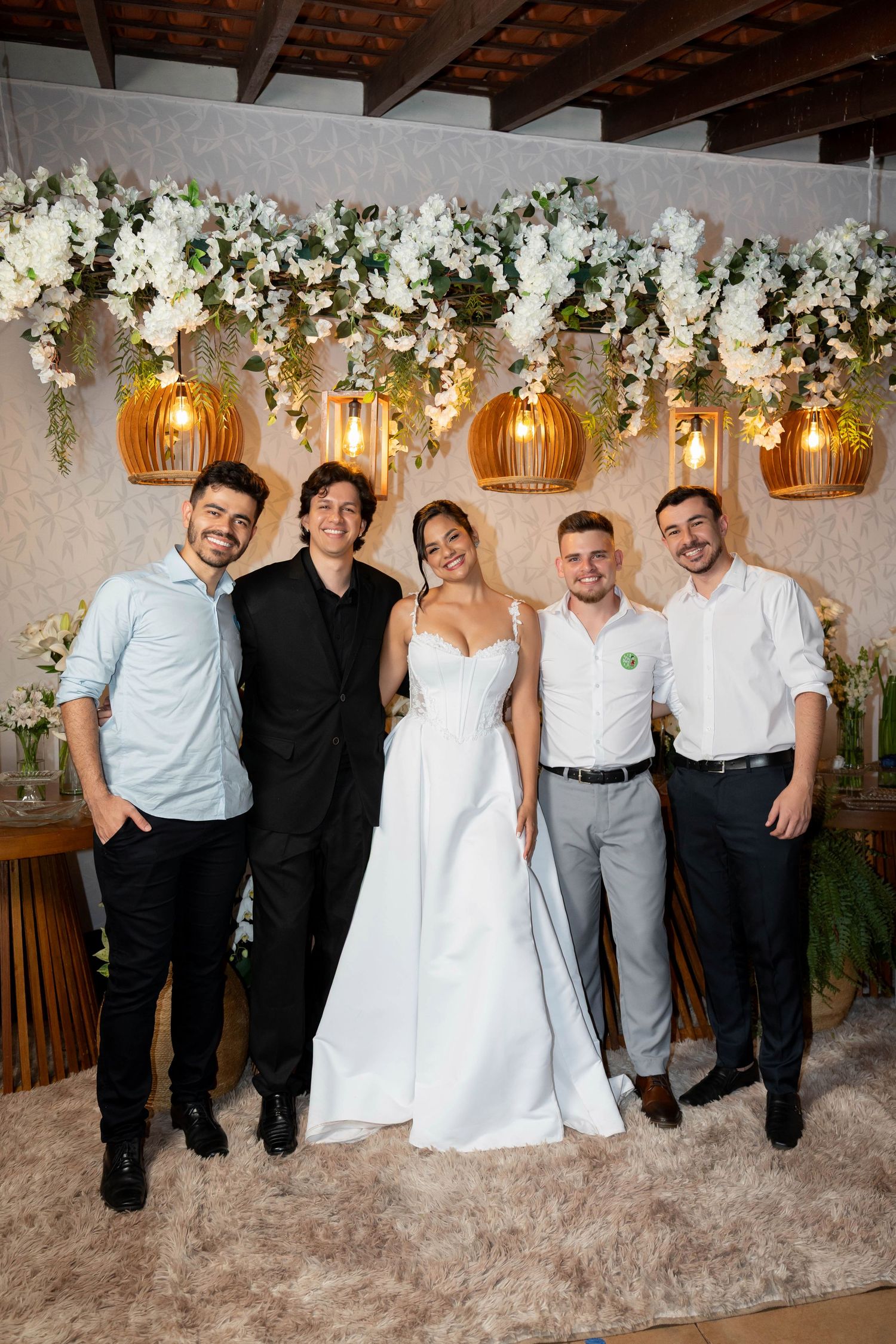 Bride in white gown poses with four guests at elegant wedding reception with floral backdrop and warm pendant lights.