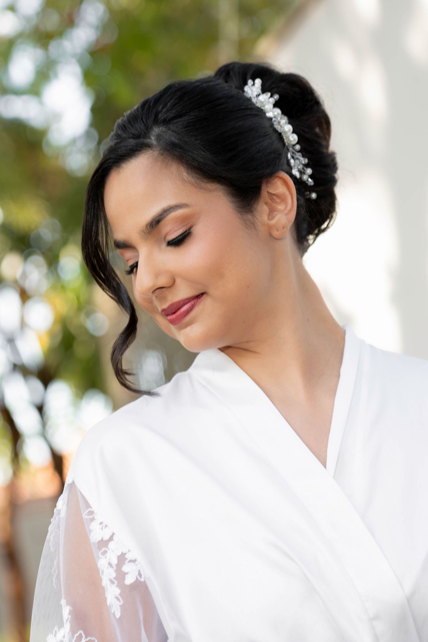 A bride in a white robe with floral hair accessory smiles softly, wearing elegant bridal makeup.