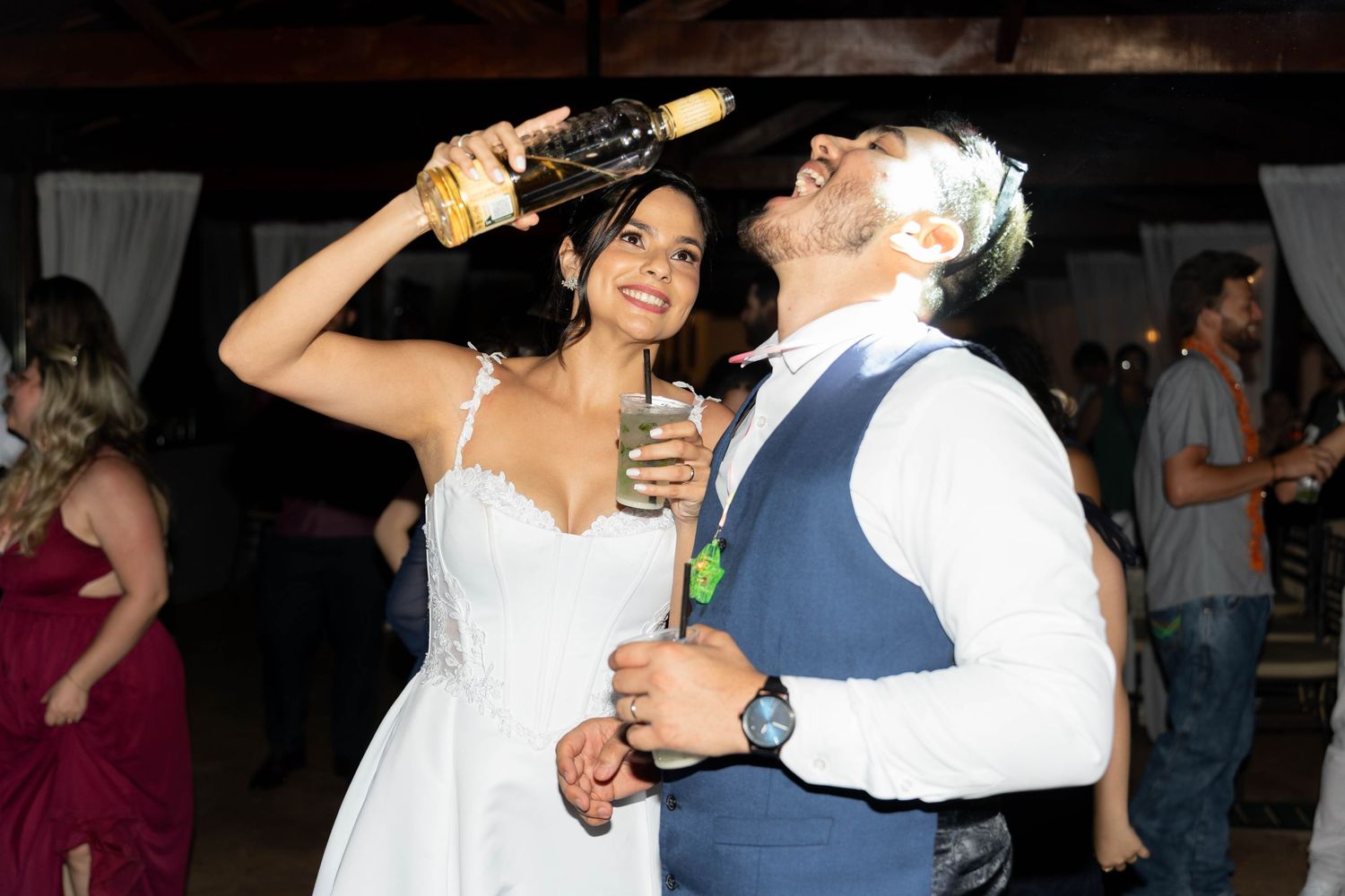 Bride in white dress pours champagne into groom's mouth on wedding dance floor.