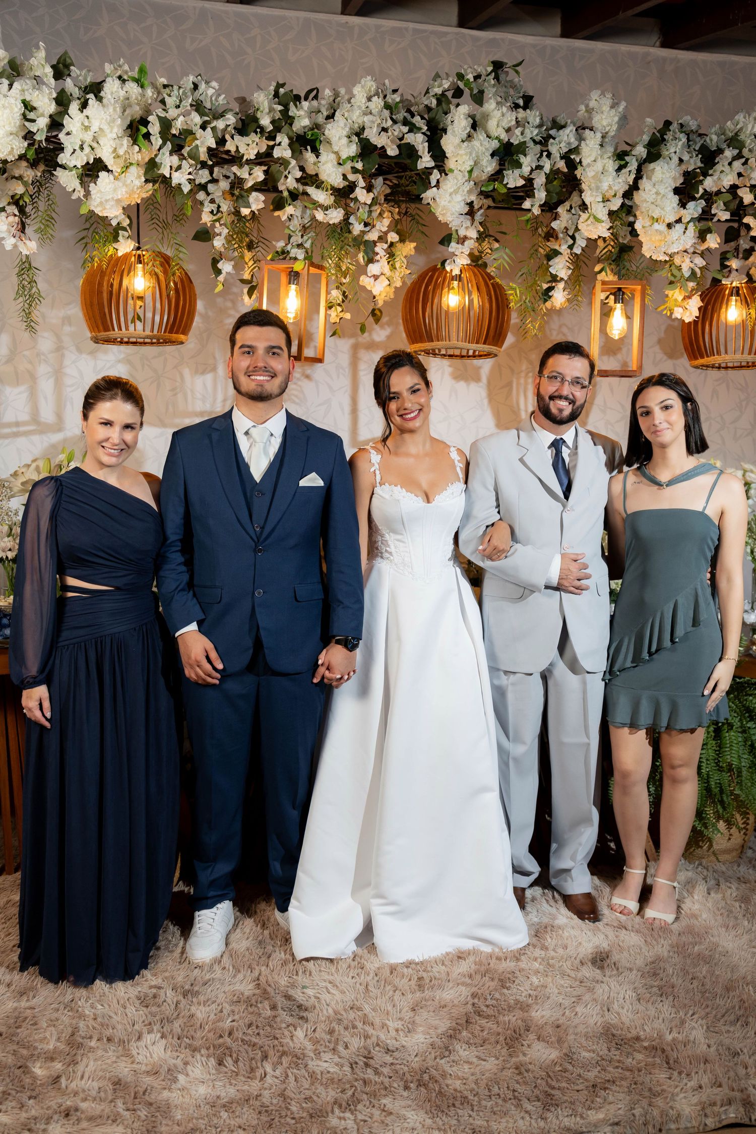 Wedding party of five posing elegantly under floral arrangements and pendant lights, bride in white gown with groomsmen and guests.