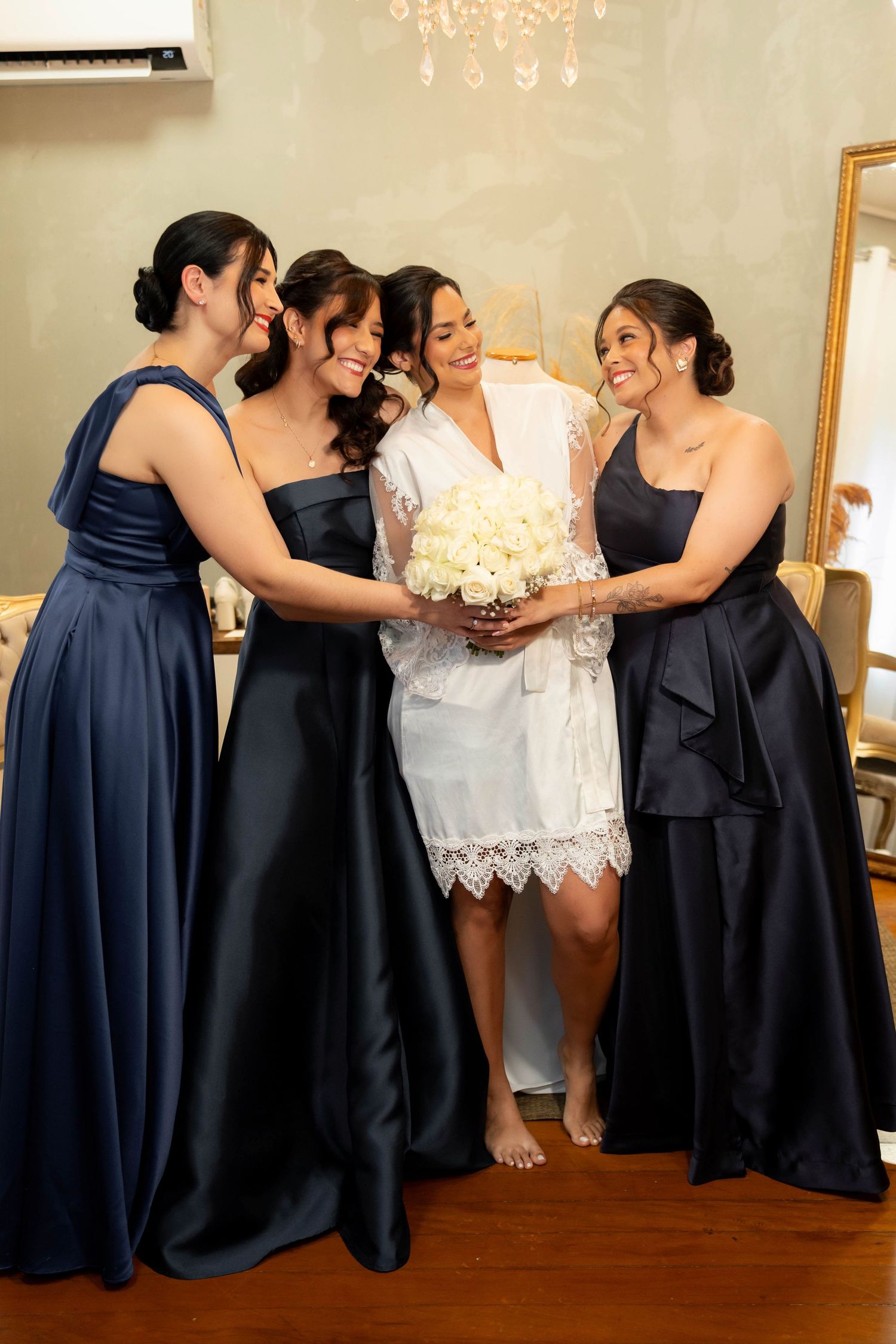 Bride in white robe holds bouquet surrounded by three bridesmaids in navy blue gowns in an elegant dressing room.