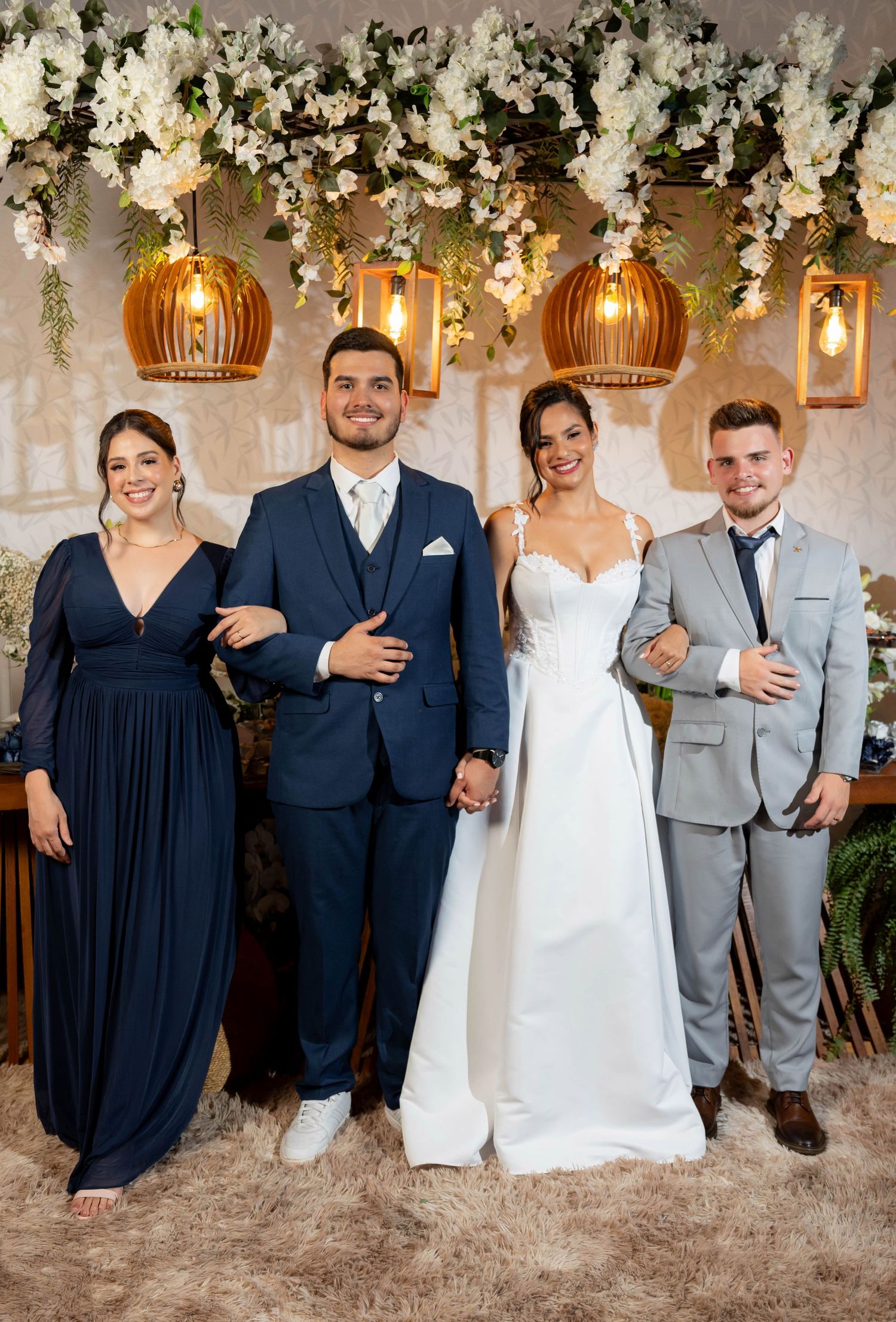 Wedding party poses under floral arch with pendant lights, bride in white gown flanked by guests in navy and gray suits.