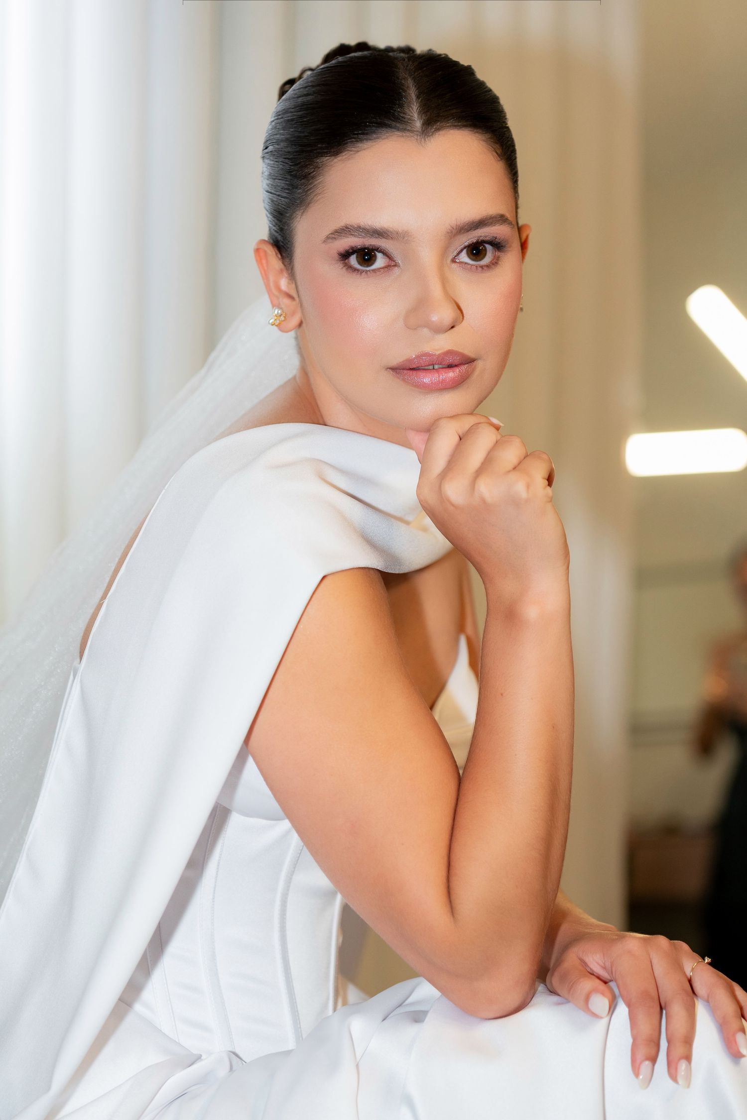 Elegant bride in white dress and veil poses gracefully, showcasing soft bridal makeup and braided updo hairstyle.