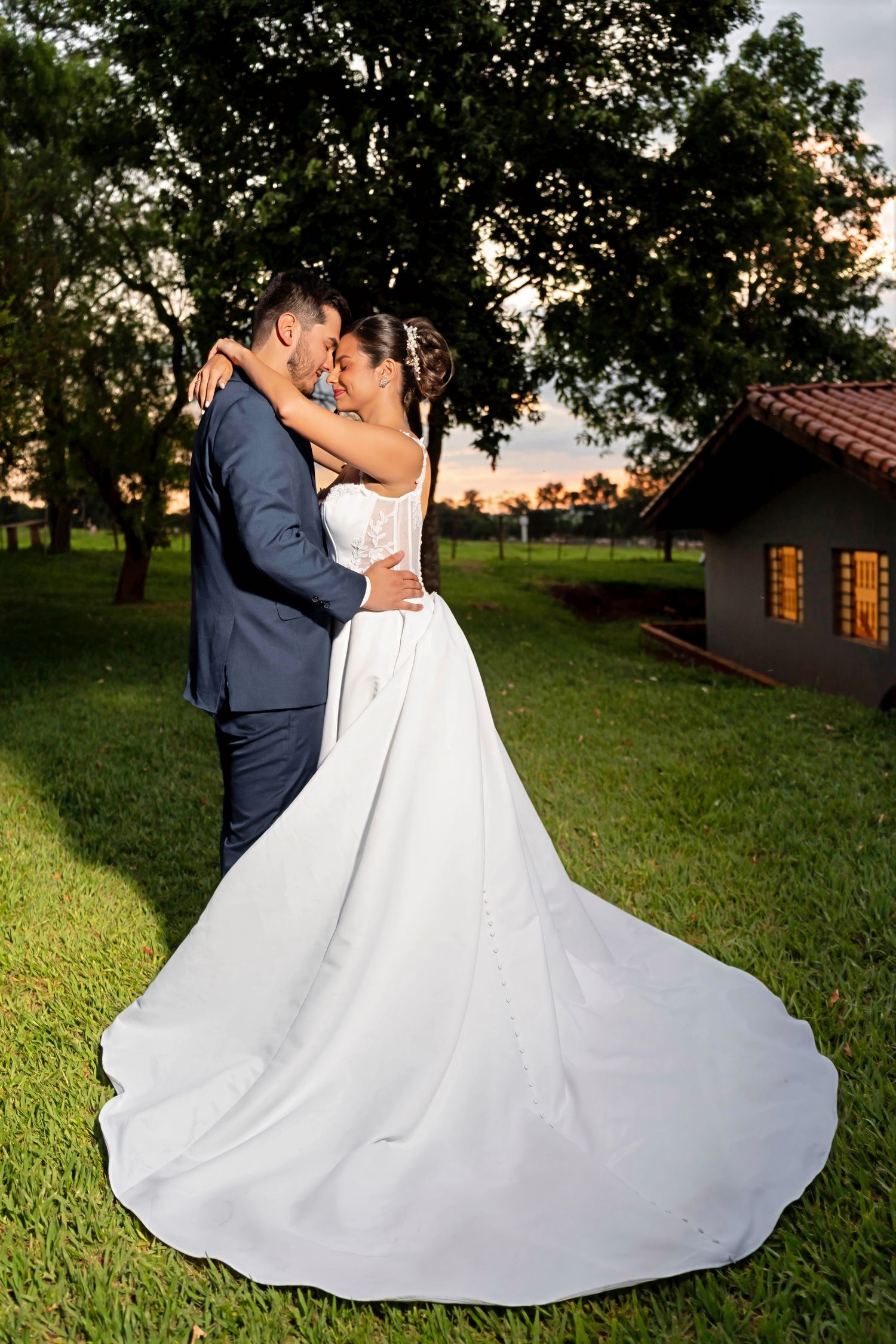 Bride and groom share a romantic kiss at sunset, with the bride's elegant white gown train spread across lush green grass.