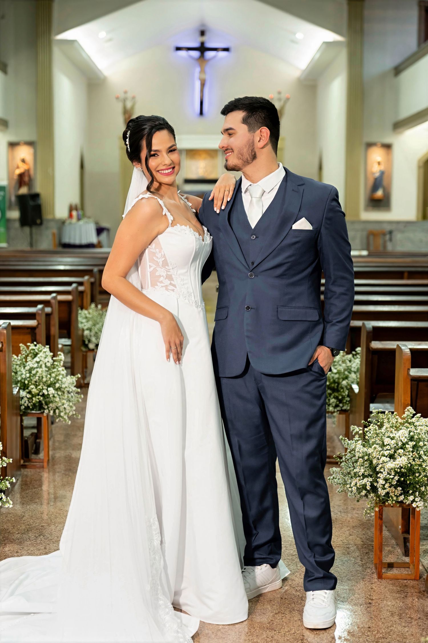 Bride in white gown and groom in navy suit pose together in a decorated church aisle on their wedding day.