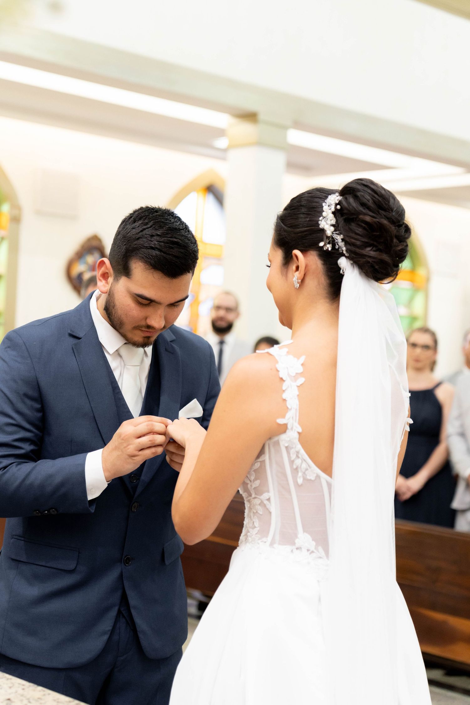 Groom in blue suit placing wedding ring on bride's finger during church ceremony.