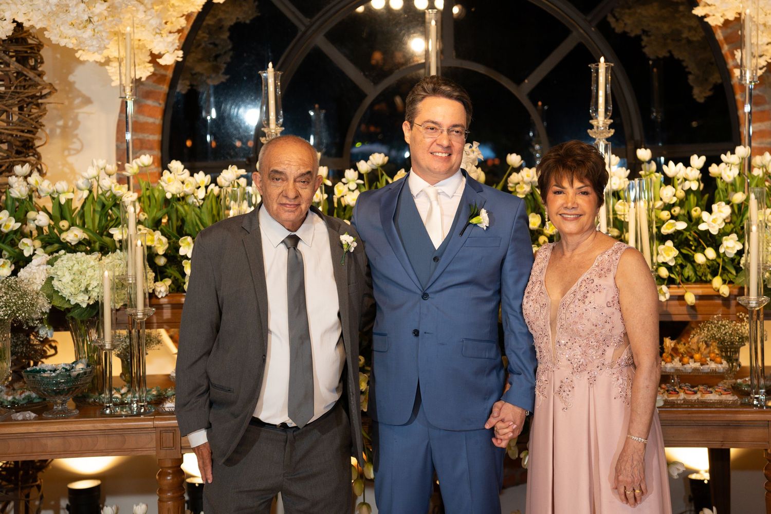 Groom in blue suit poses with two guests at elegant floral wedding reception.