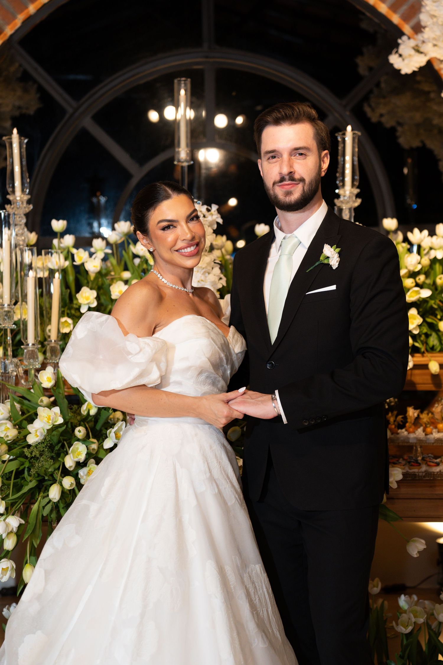 A bride in a white puffed-sleeve gown and groom in a black tuxedo pose together at an elegant floral wedding venue.