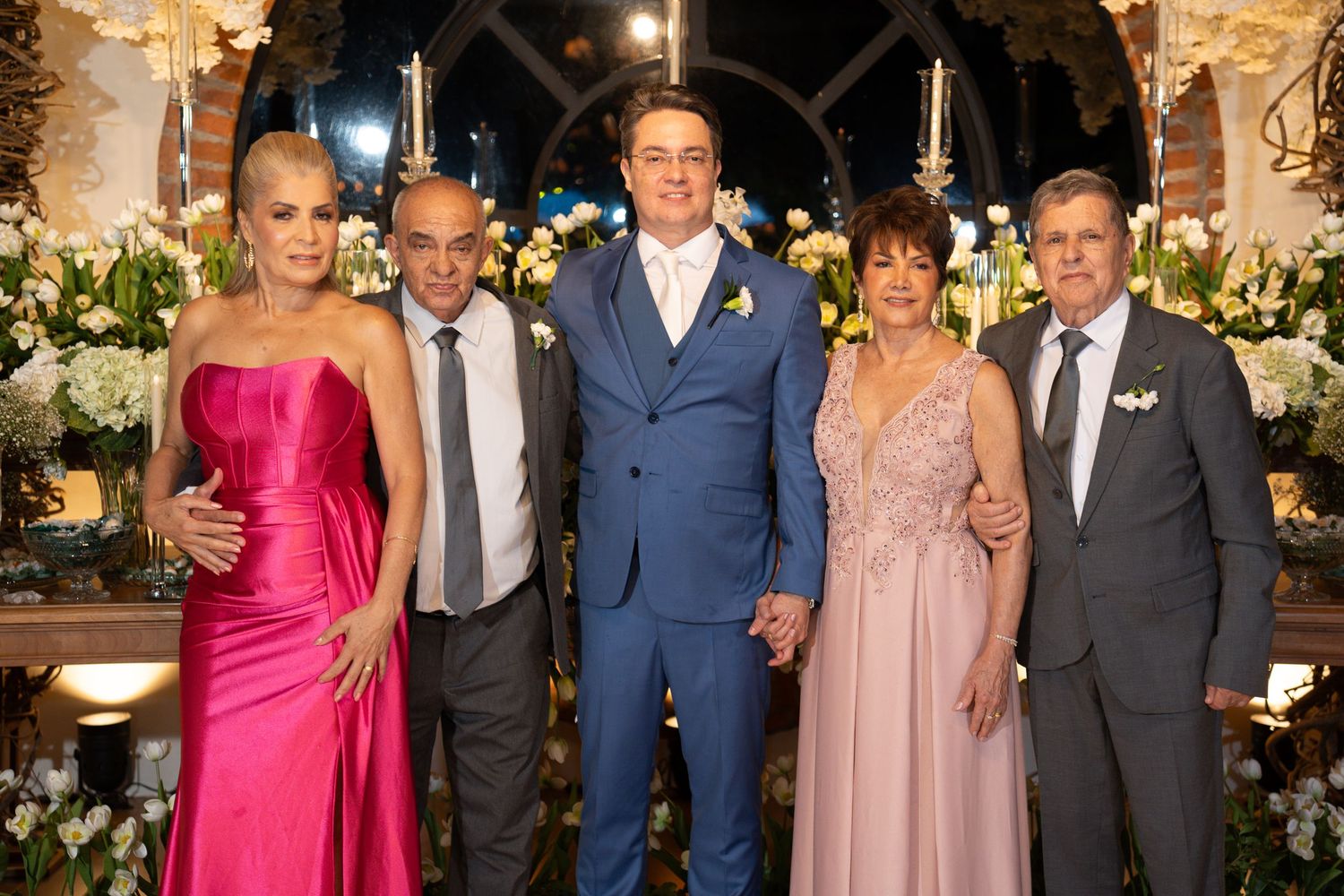 Five elegantly dressed people pose together at a formal wedding reception with floral decorations.