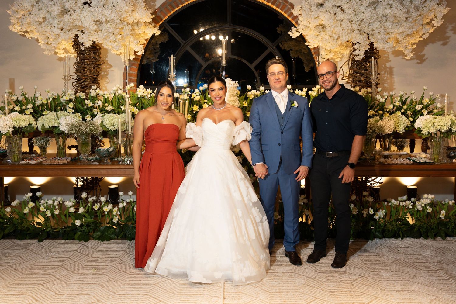 Elegant wedding reception with bride in white gown, groom in blue suit, and guests in formal attire amid floral decor.