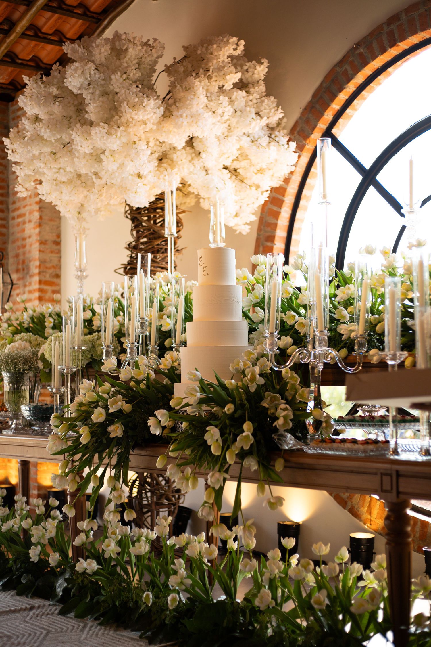 Elegant wedding cake display with white floral arrangements, candelabras, and lush greenery in a rustic venue.
