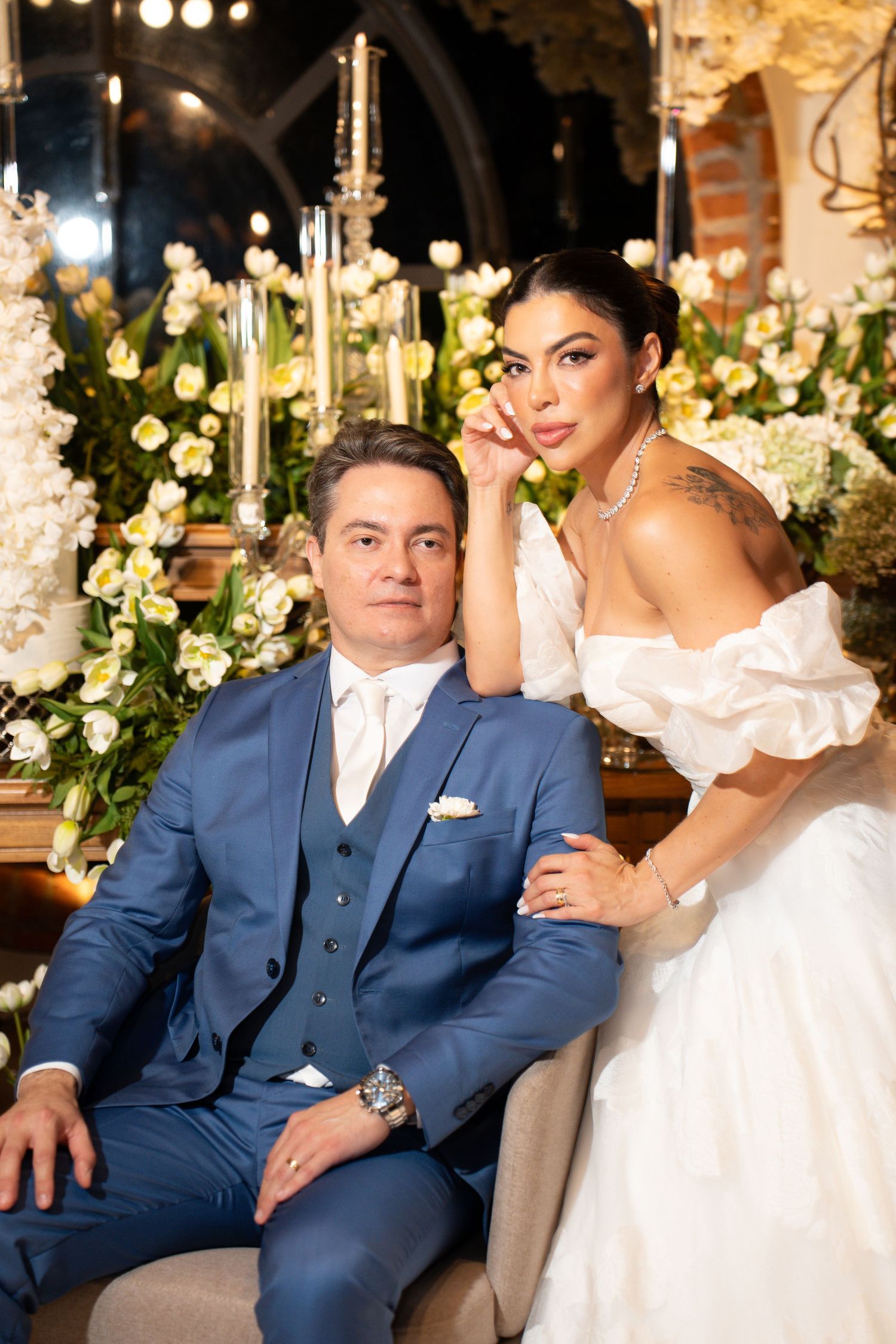 Elegant wedding couple posing together, groom in blue suit and bride in white off-shoulder gown, surrounded by floral arrangements.