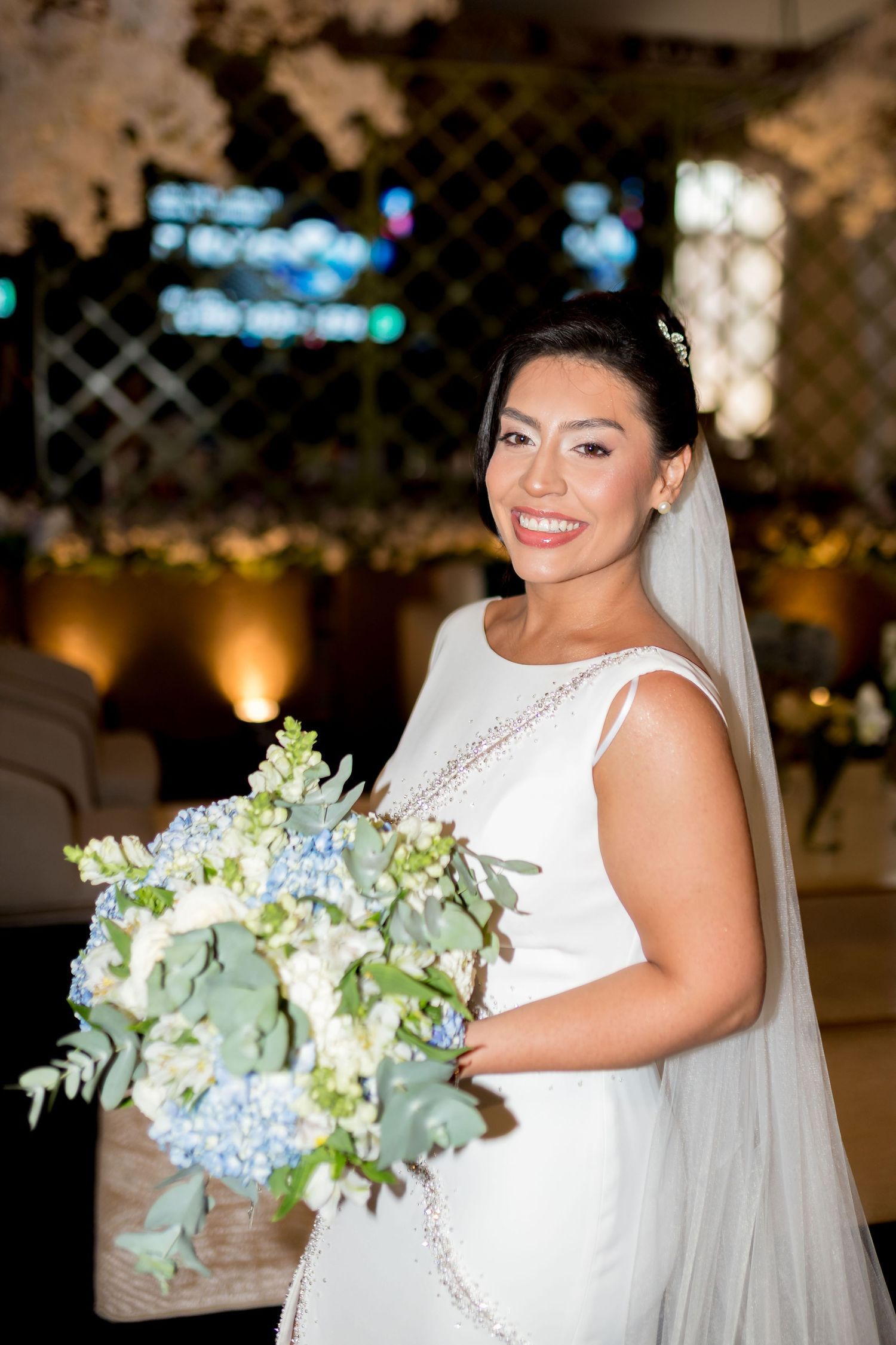 Smiling bride in white gown holding blue hydrangea bouquet with greenery, wearing veil at elegant wedding venue.
