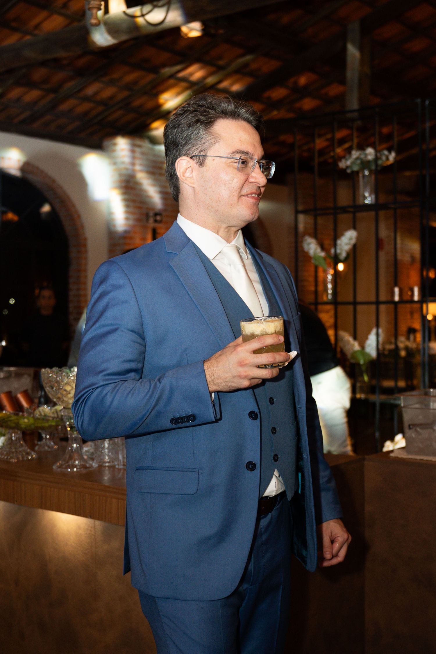 Man in blue three-piece suit holding a drink at an elegant indoor event venue.