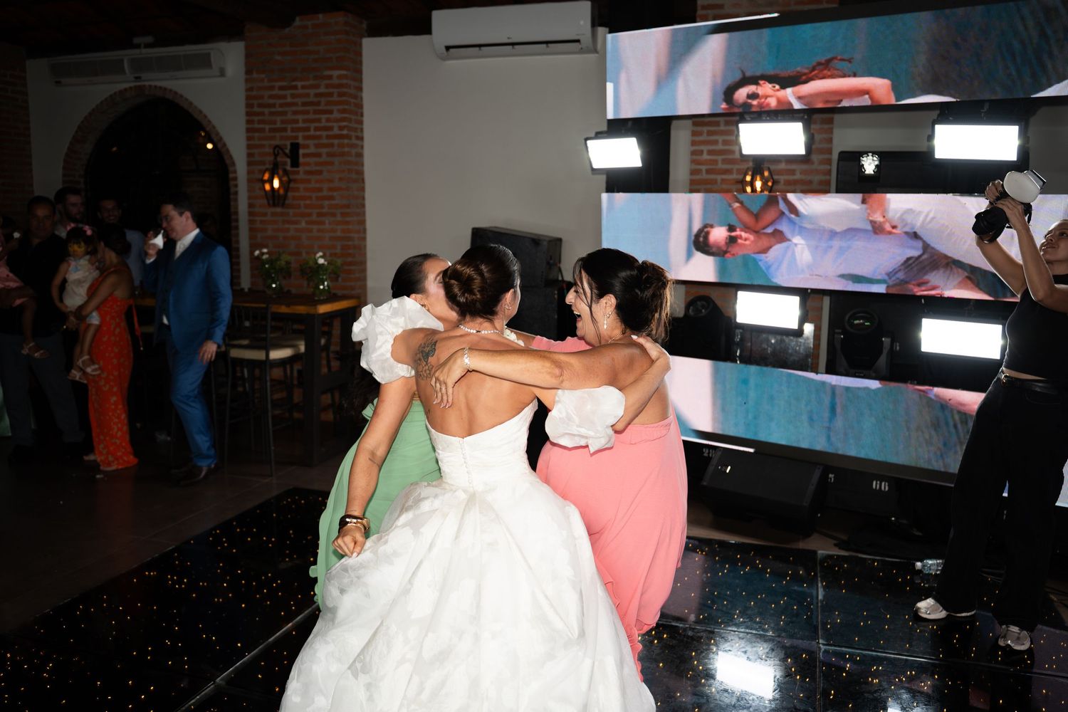 Bride in white gown embraced by bridesmaids on wedding dance floor with large screen display in background.