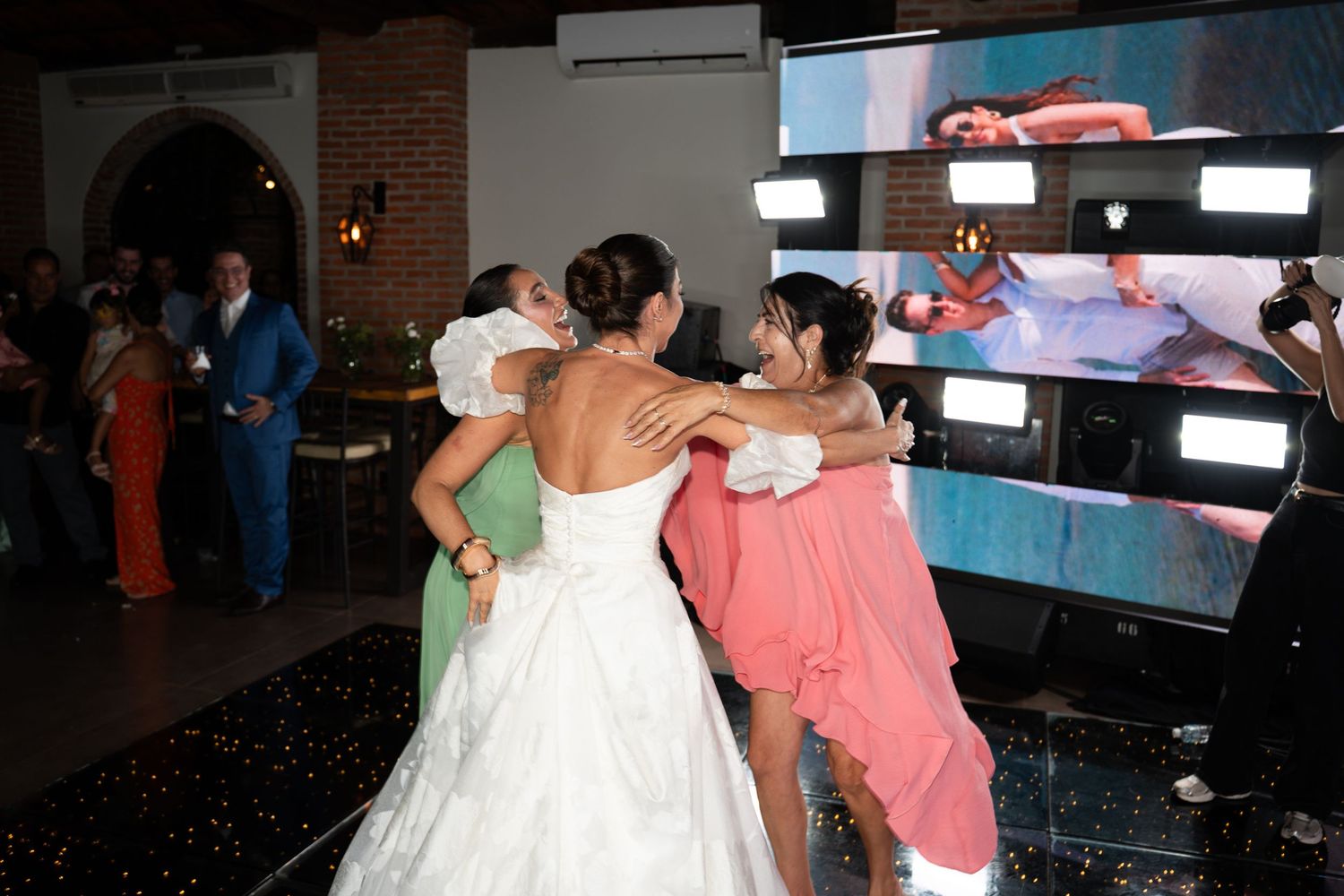 Bride in white gown embraced by bridesmaids in coral and green dresses on a dance floor at a wedding reception.