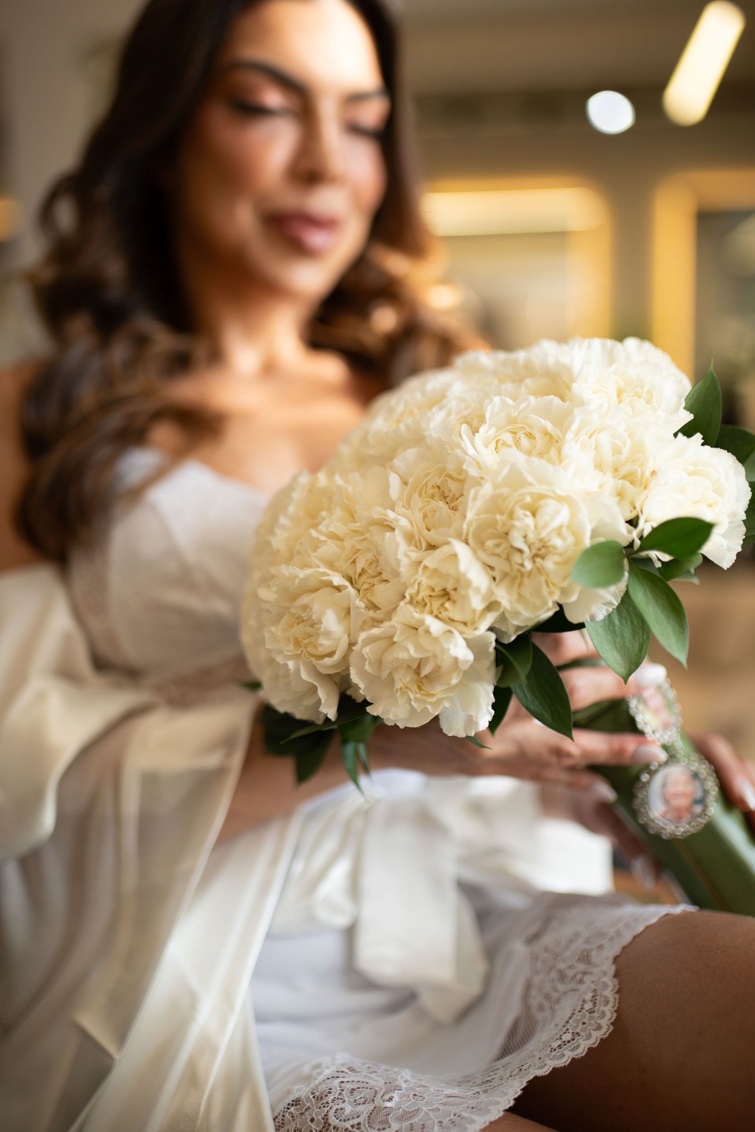 Close-up of bridal bouquet of white carnations with ribbon, held by bride in white lace and satin robe.