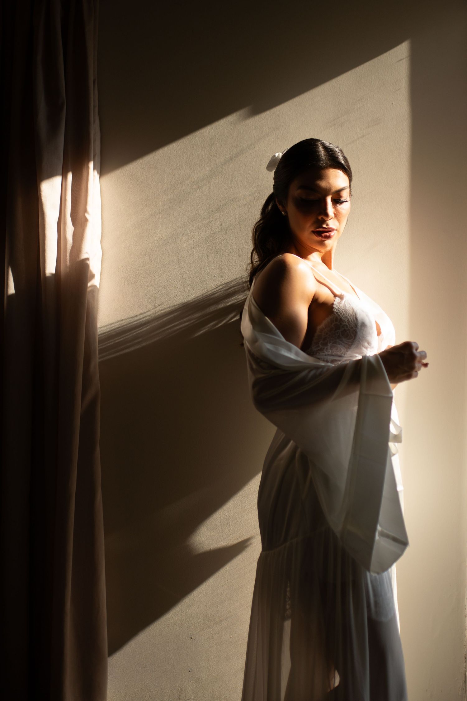 A bride in a white lace gown and sheer wrap stands in dramatic sunlight near sheer curtains.