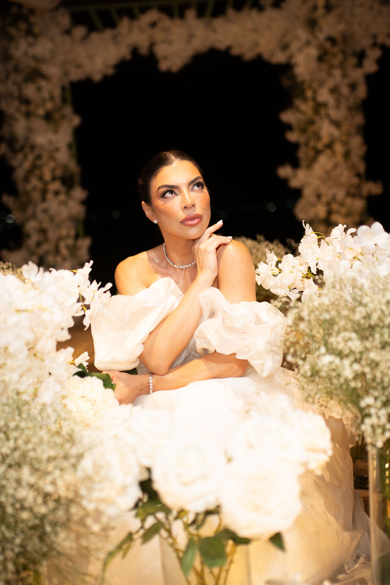 Elegant bride in white off-shoulder gown surrounded by white floral arrangements, gazing upward dreamily.