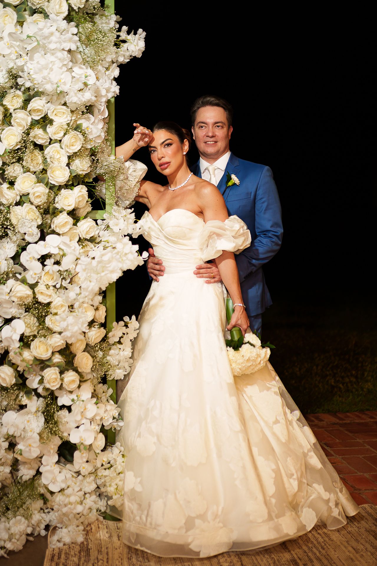 Elegant bride in strapless gown and groom in blue suit pose beside white floral arch at nighttime wedding ceremony.