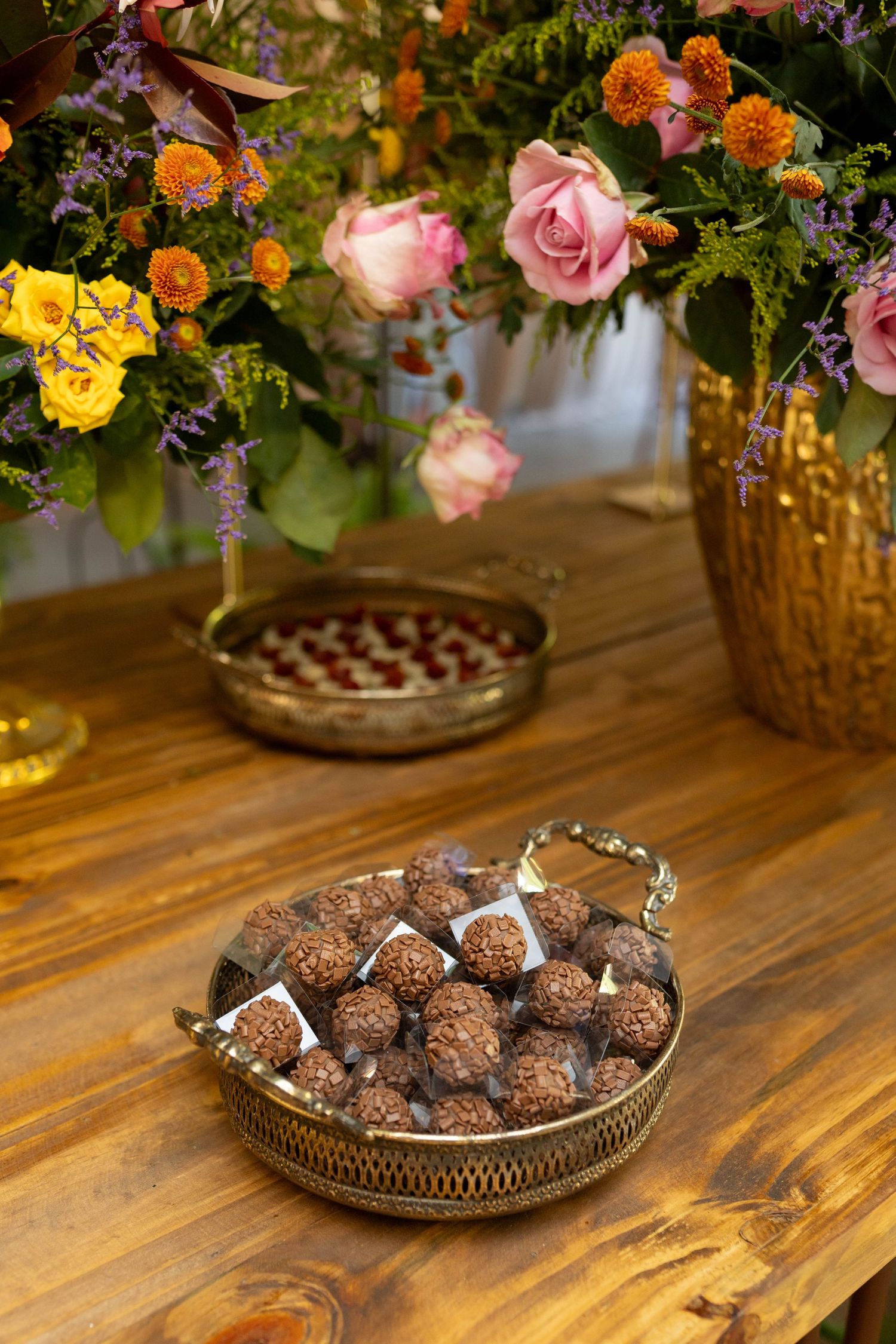 Gold tray of chocolate truffles on wooden table with floral arrangement of pink roses and orange blooms in background.