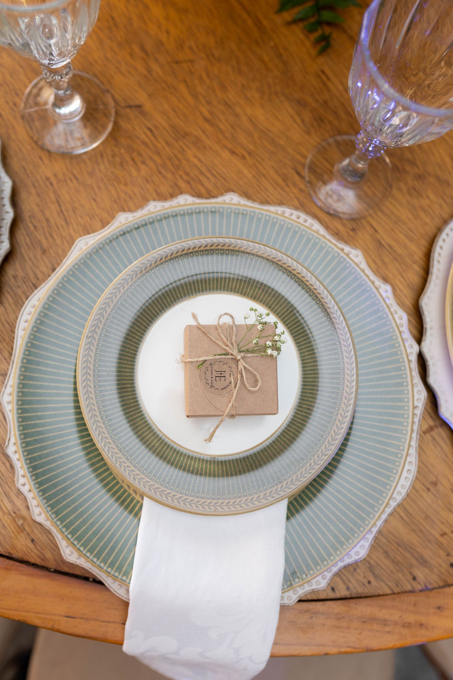Elegant wedding place setting with sage green plates, white napkin, and kraft paper favor box tied with twine on wooden table.