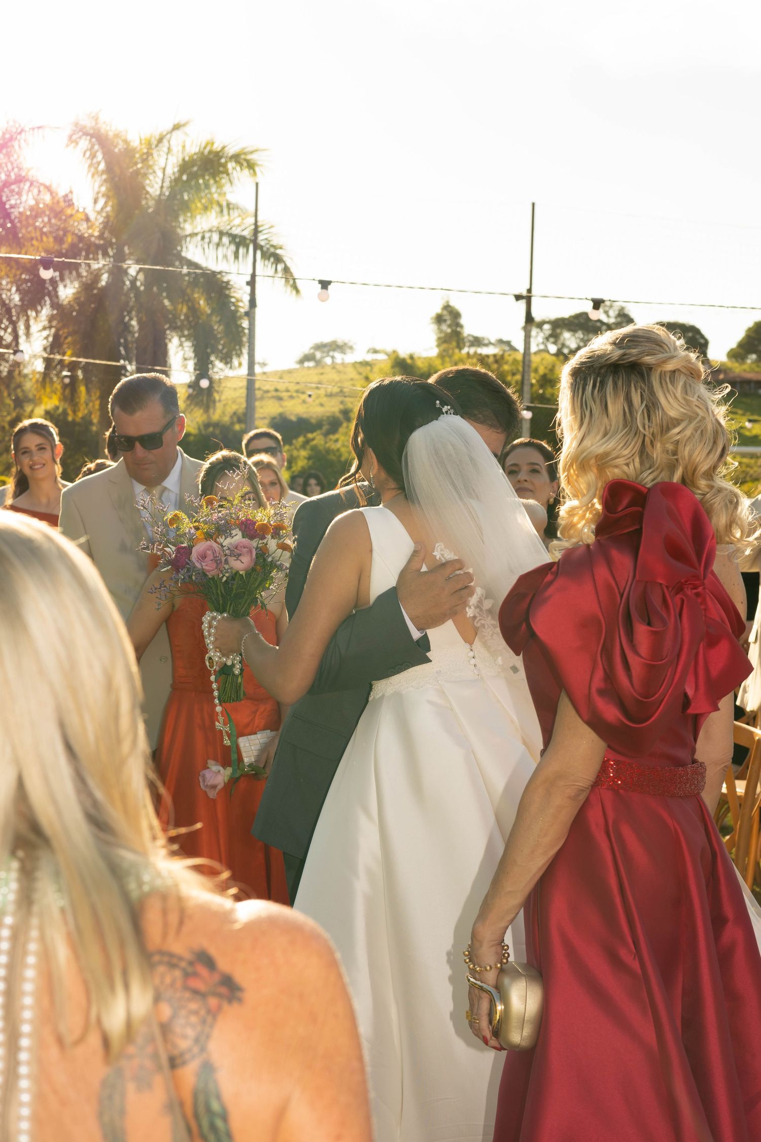 Bride and groom share a kiss at outdoor wedding ceremony surrounded by guests in red dresses and warm golden sunlight.