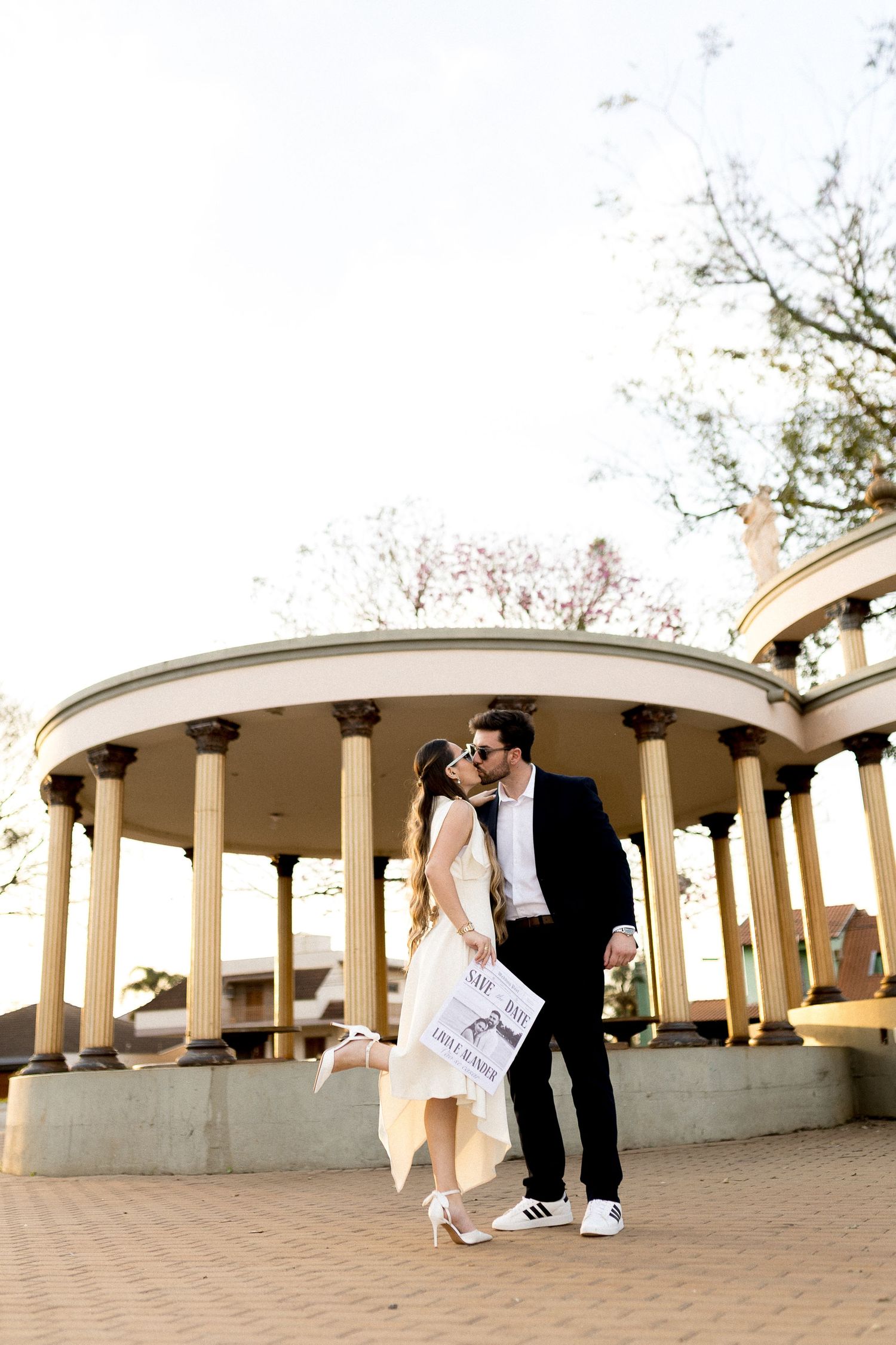 Couple kissing in front of a elegant columned rotunda, man in black suit, woman in white dress holding a sign.