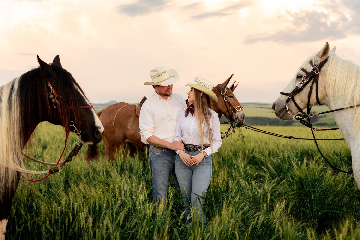 Couple in cowboy hats standing romantically among horses in a green field at sunset.