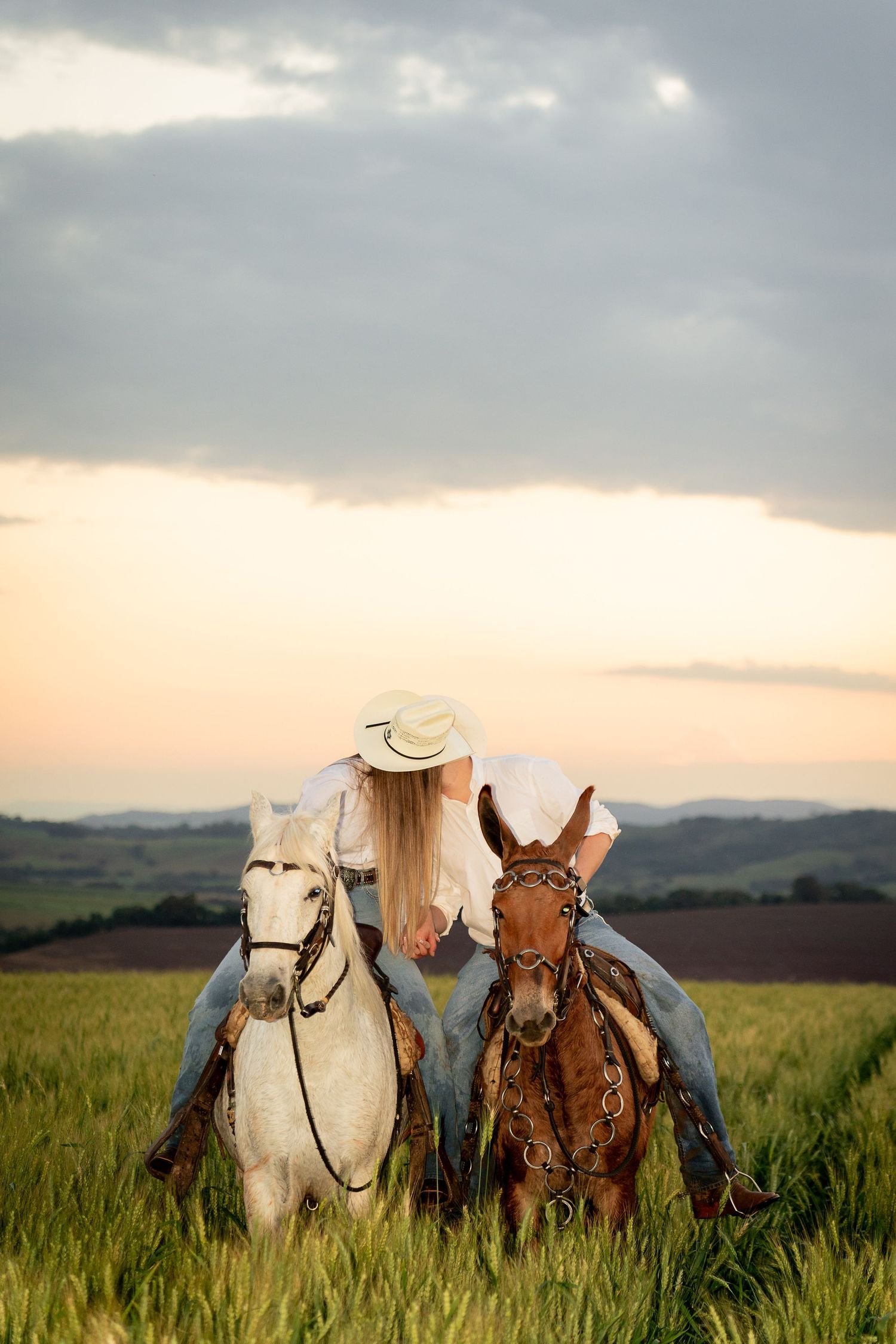 Two women on horseback riding through a green field at sunset with rolling hills in the background.