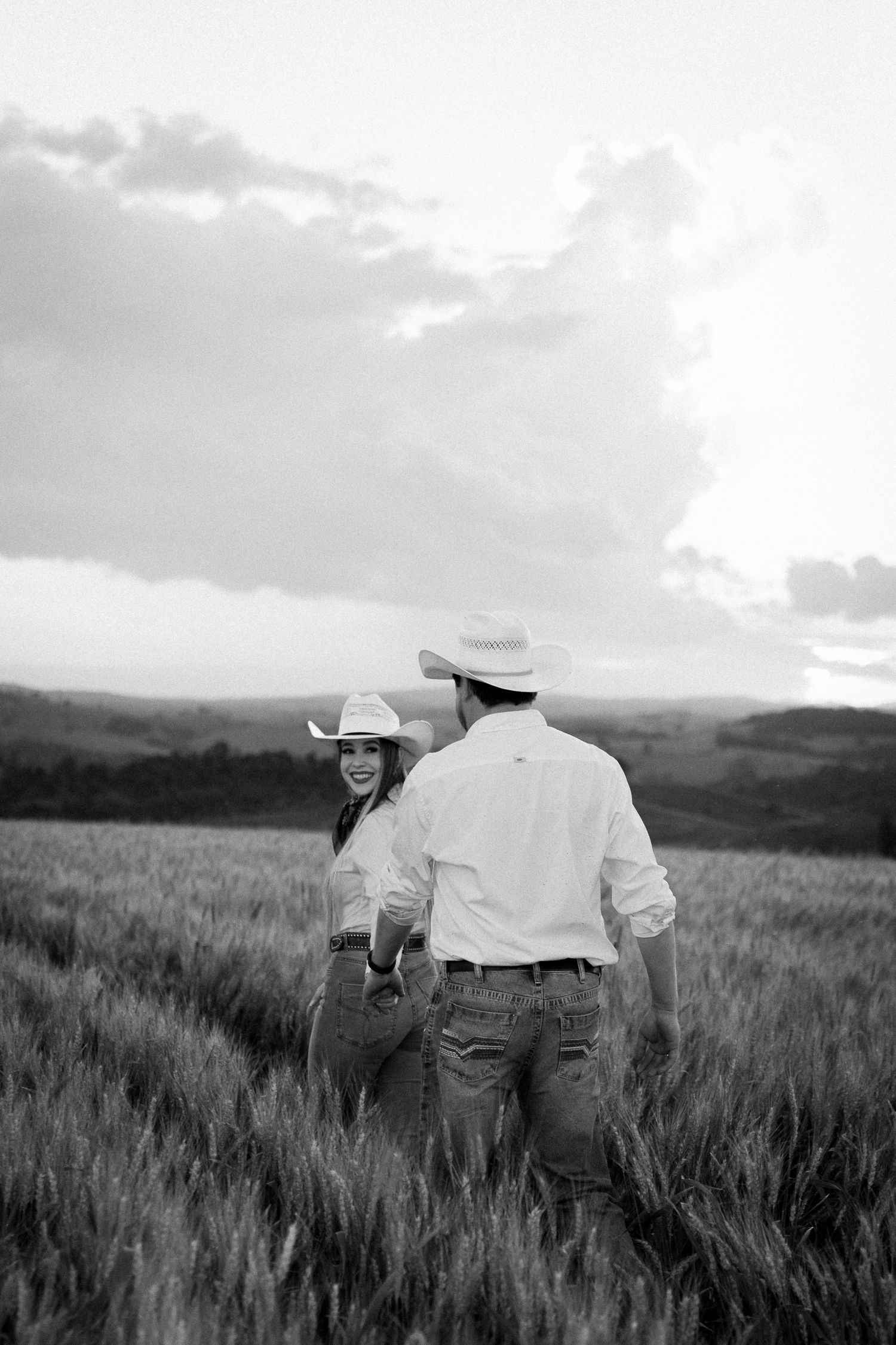 Black and white photo of a couple in cowboy hats holding hands in a wheat field at sunset.