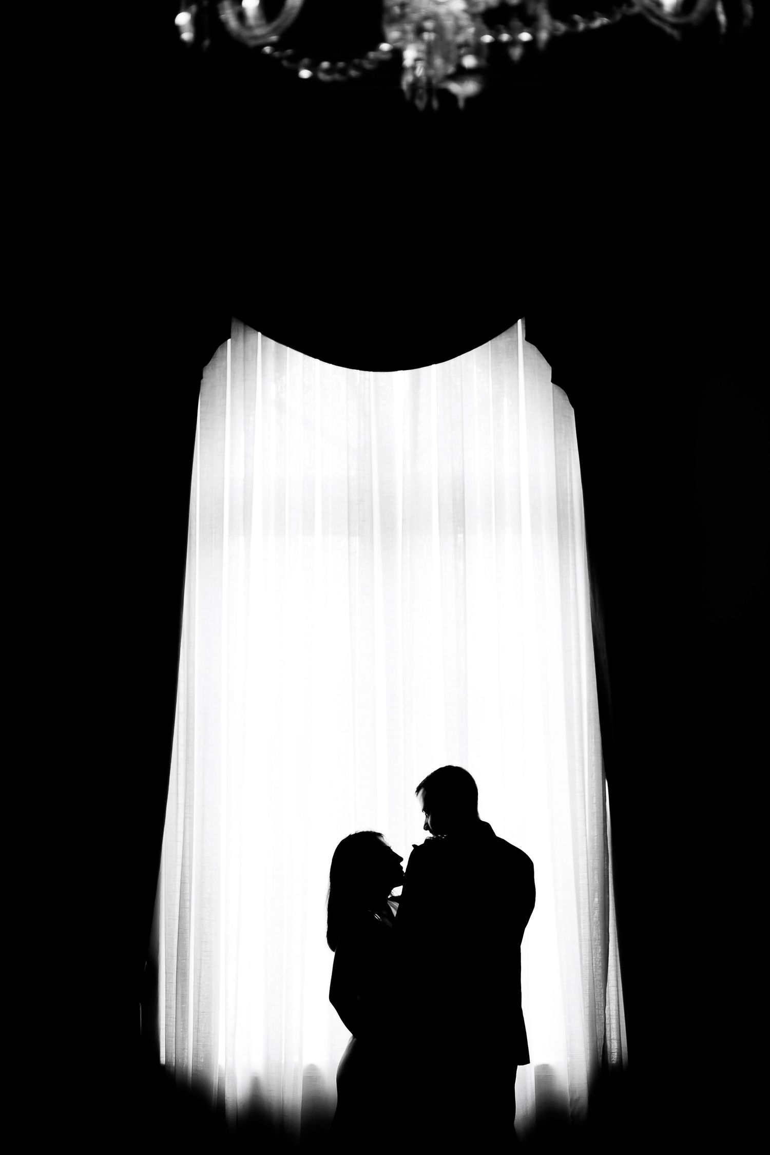 Romantic black and white silhouette of a couple standing before a glowing curtained window with chandelier above.