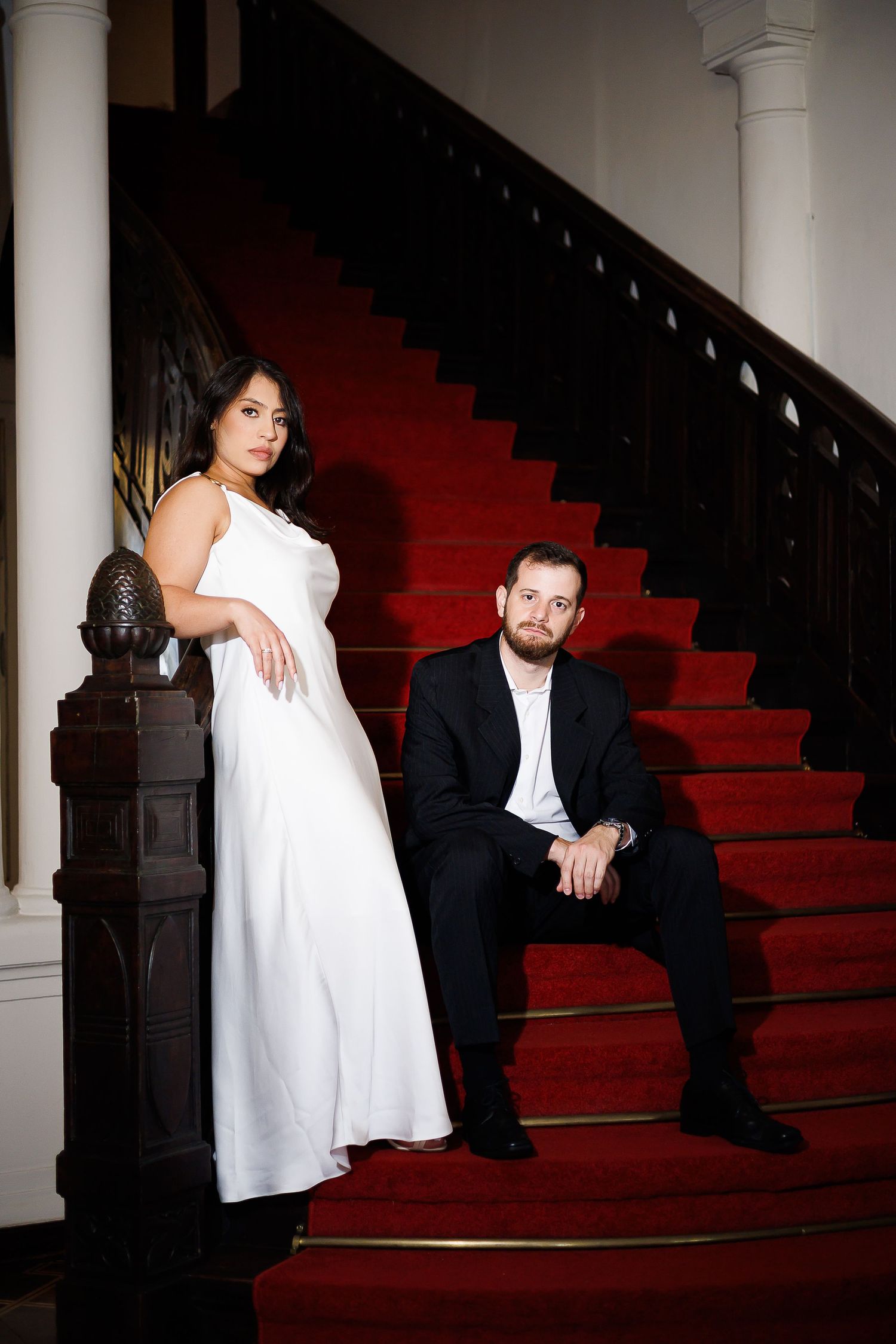 Elegant couple posing on red carpeted grand staircase, woman in white gown standing, man in black suit seated.