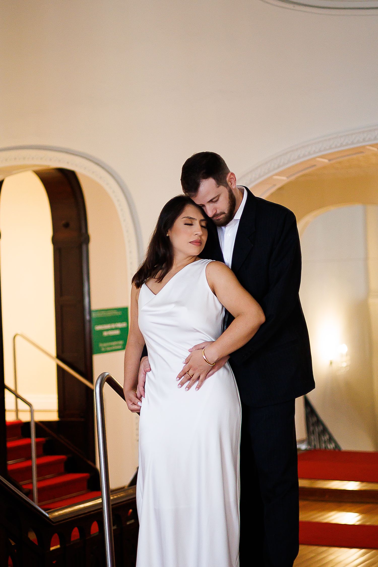 Couple in elegant wedding attire embrace on red-carpeted stairs, bride in white gown, groom in black suit.