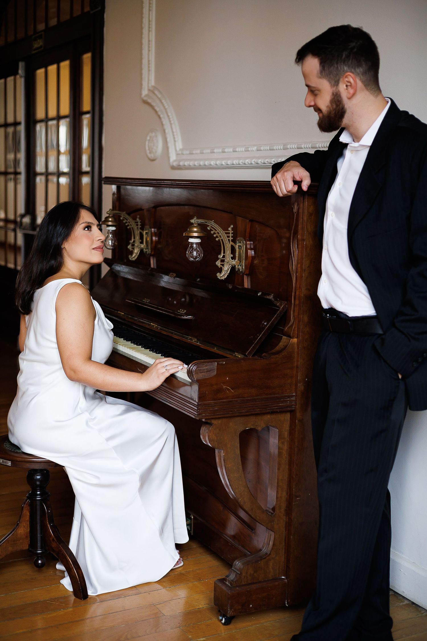 Woman in white dress plays upright piano while man in tuxedo leans on it, sharing an elegant musical moment.
