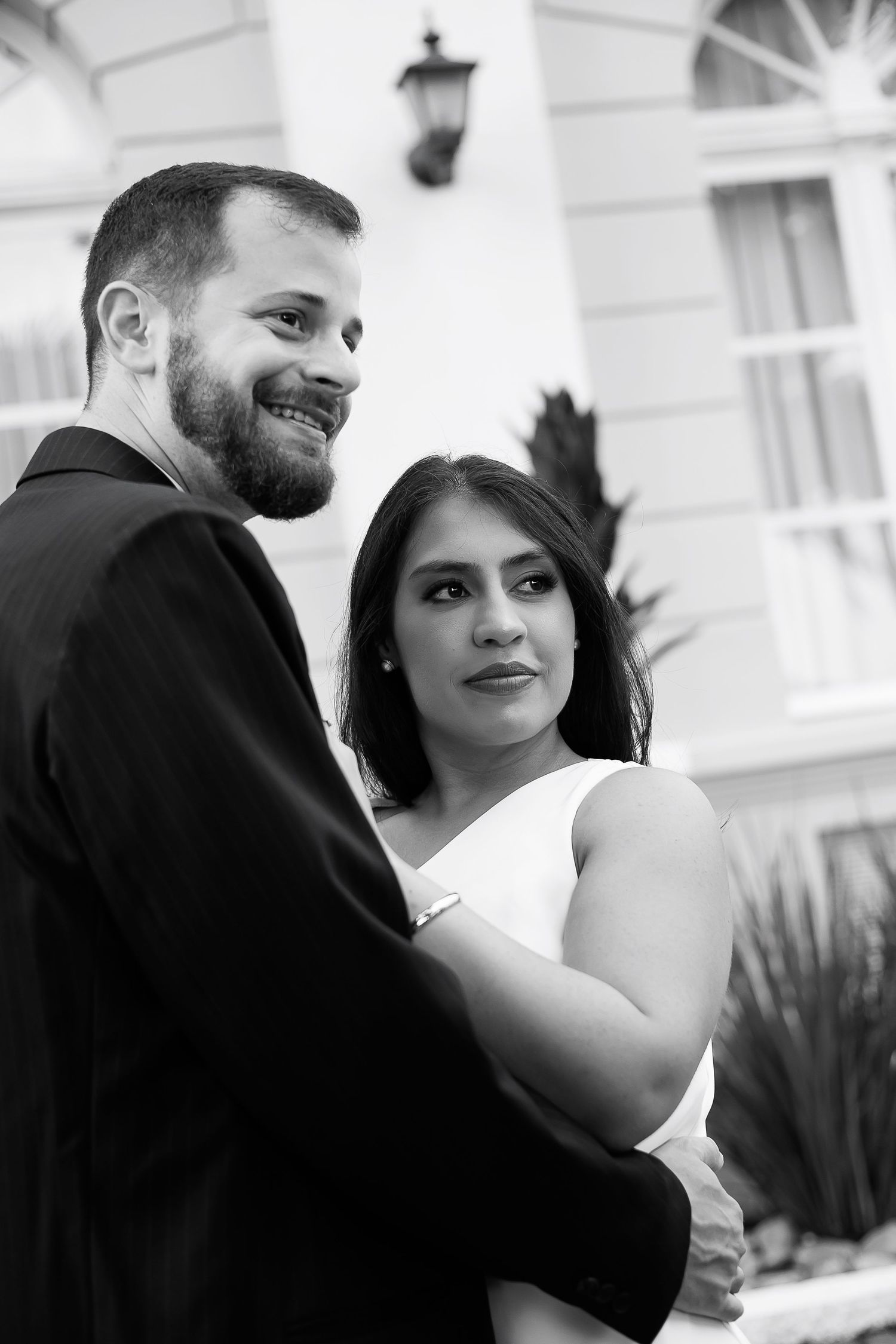 Black and white wedding photo of smiling groom and bride embracing outdoors near elegant white building.